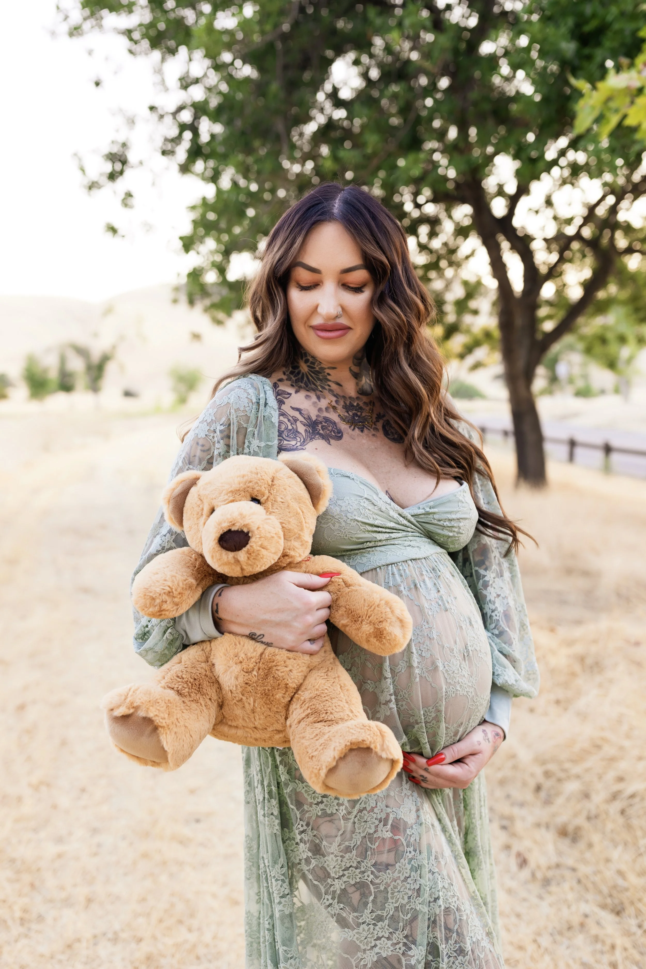 A pregnant woman with long wavy brown hair, tattoos on her neck and hands, wearing lace clothing, holding a teddy bear in an outdoor park setting with trees and a pathway.