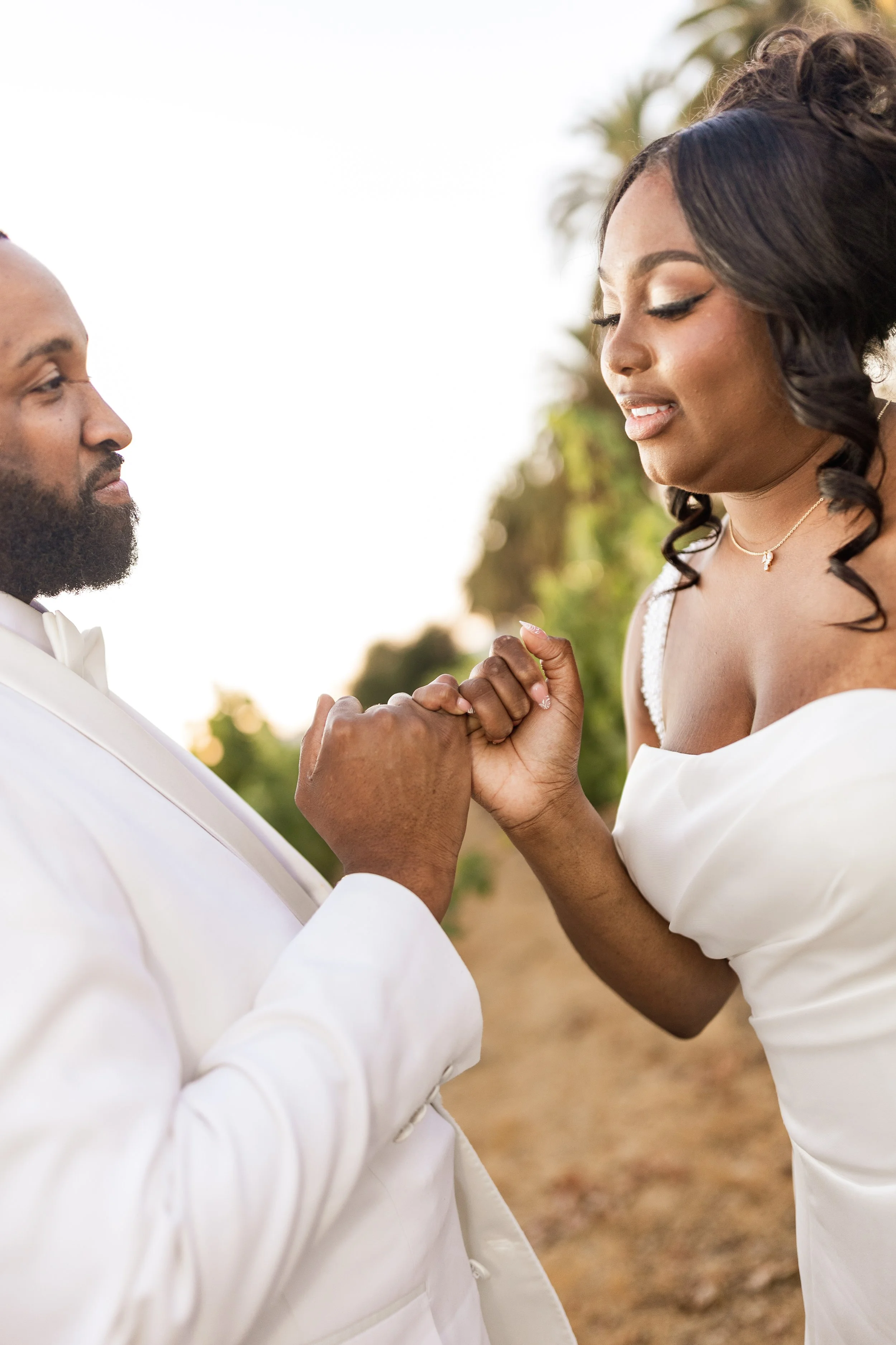 A man and woman in white suits holding hands and looking at each other outdoors with greenery in the background.