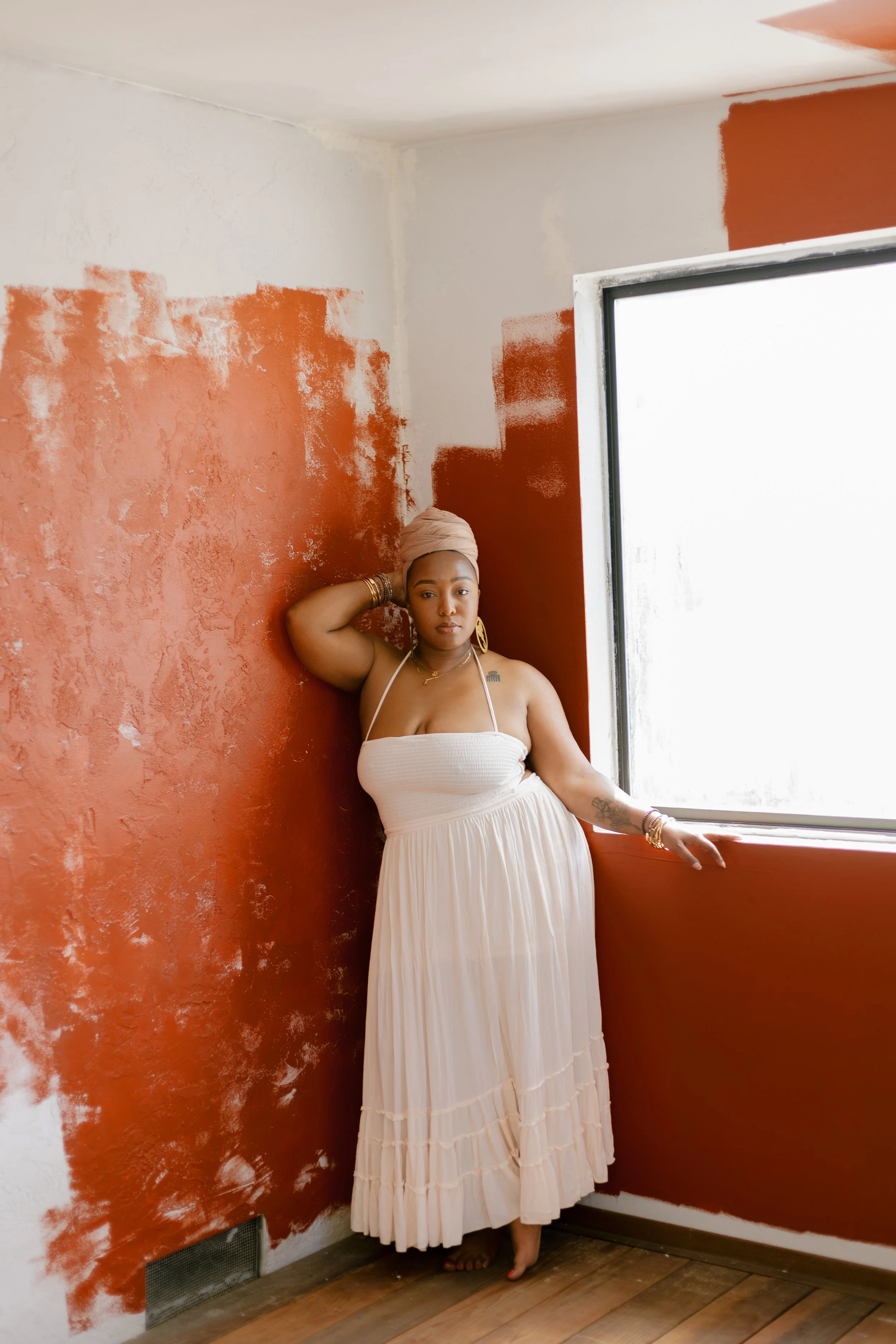 A woman in a white strapless dress with a beige headwrap and gold jewelry stands barefoot in a room with one wall painted terracotta and the other wall partially painted terracotta, near a window with white trim.