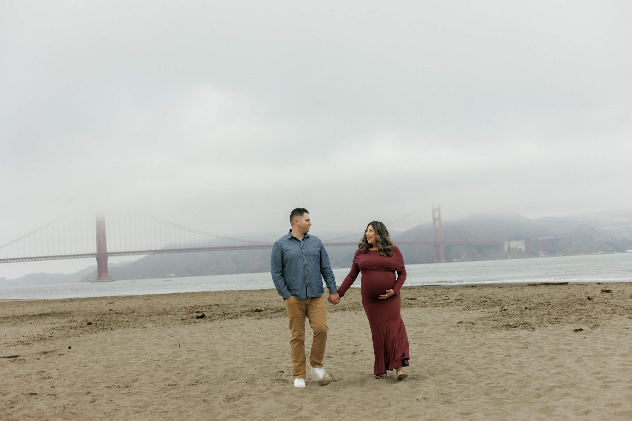 A pregnant woman and a man holding hands on a beach with the Golden Gate Bridge in the background on a cloudy day