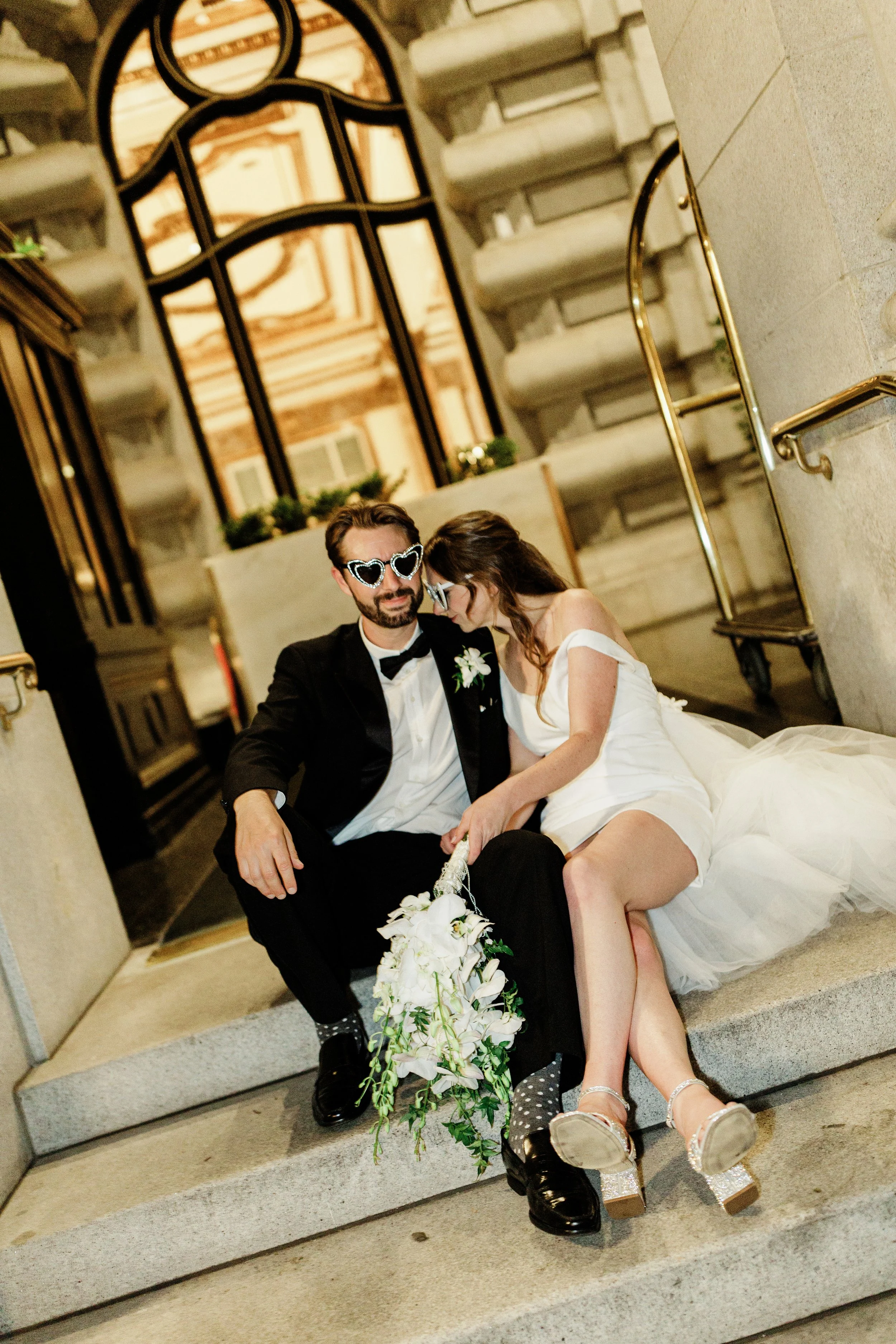 A newlywed couple sitting on steps inside a grand, elegant building. The groom is wearing a tuxedo with heart-shaped sunglasses, and the bride is in a white wedding dress, leaning on her groom. Both are smiling and holding a floral bouquet. The backg
