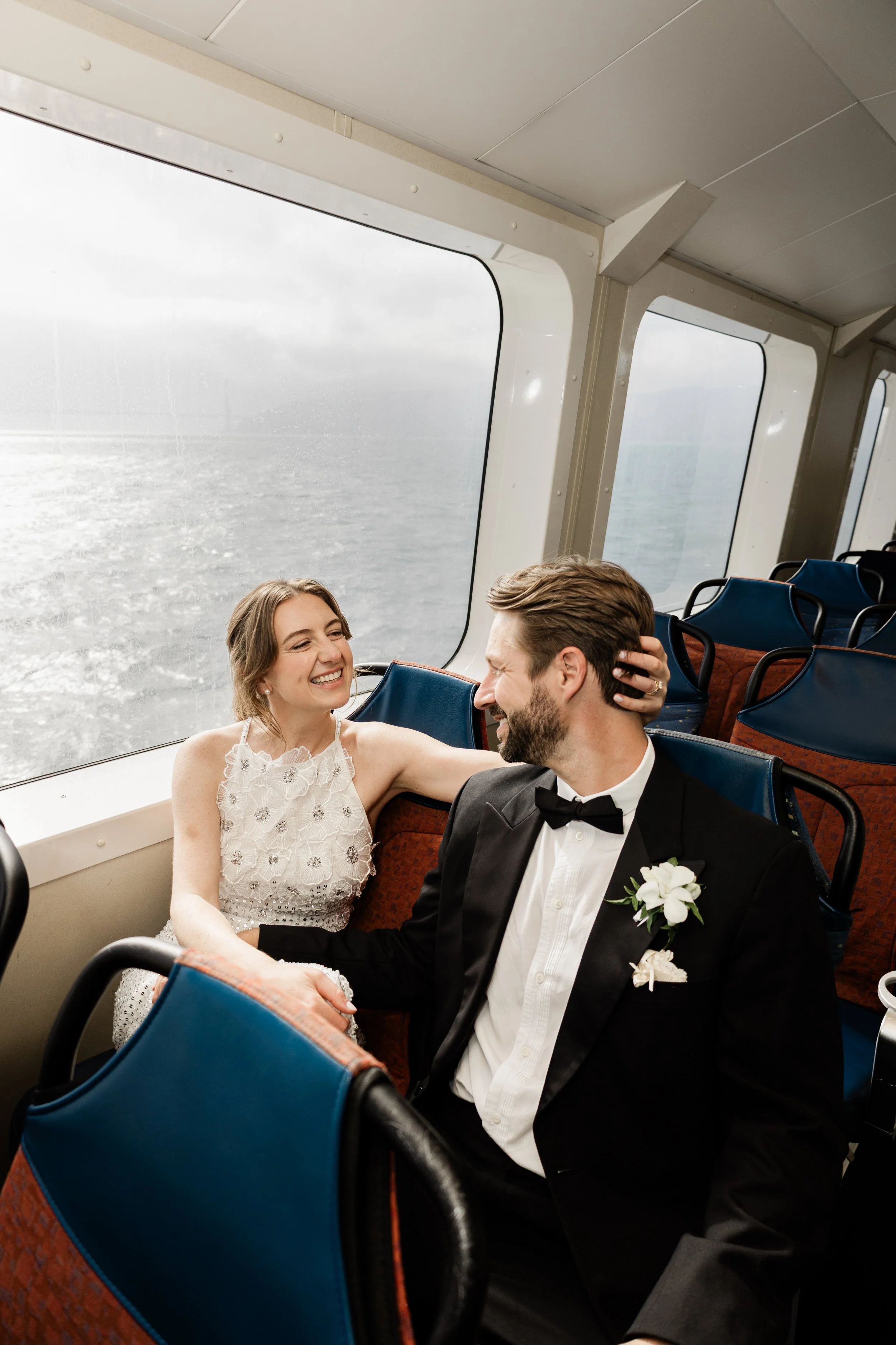 A happy couple dressed in wedding attire sitting together on a ferry boat near large windows with ocean view.