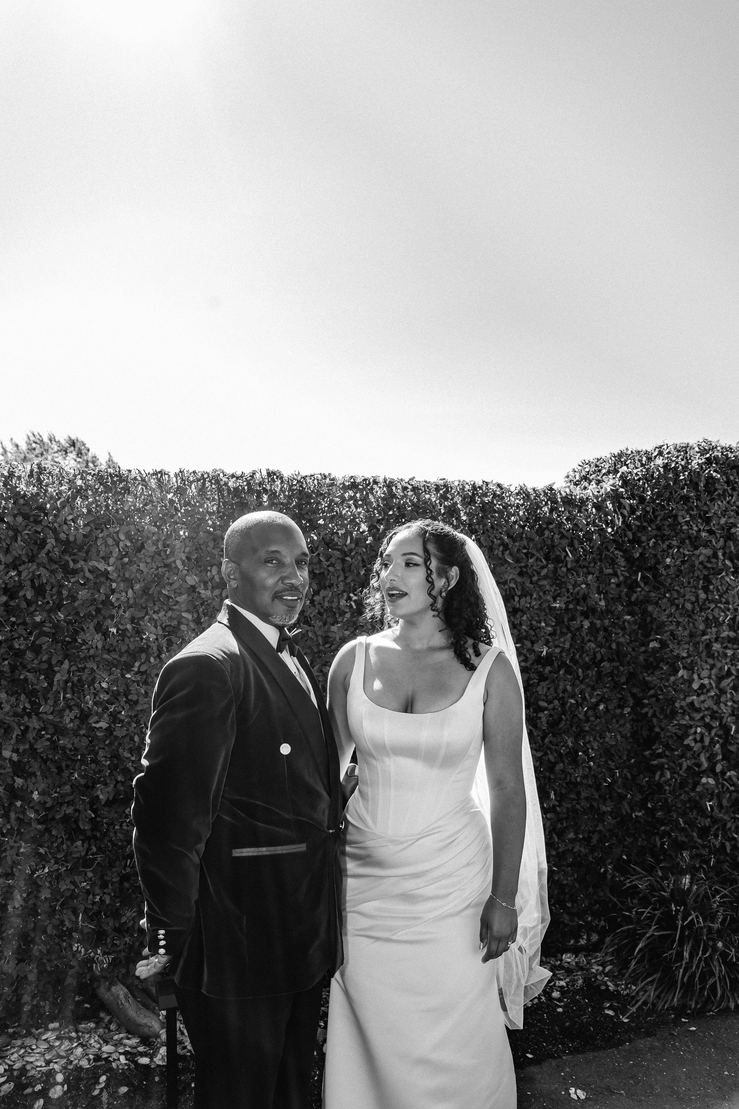 A man and woman in wedding attire standing outdoors in front of a bush. The man is wearing a tuxedo with a bowtie, and the woman is wearing a white wedding dress with a veil.