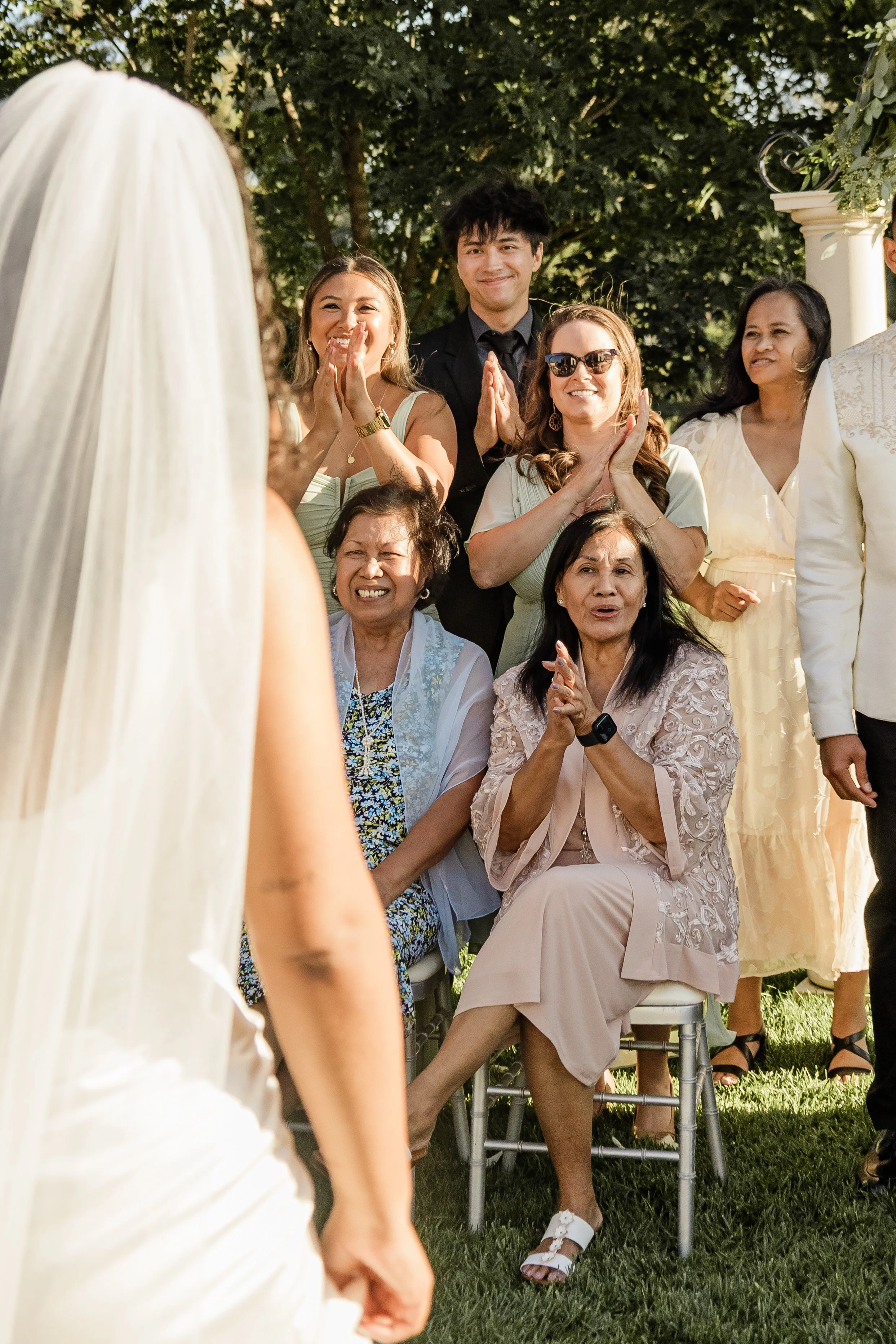 Group of women and a man at an outdoor wedding, with a woman in a wedding dress in the foreground and the guests smiling and clapping.