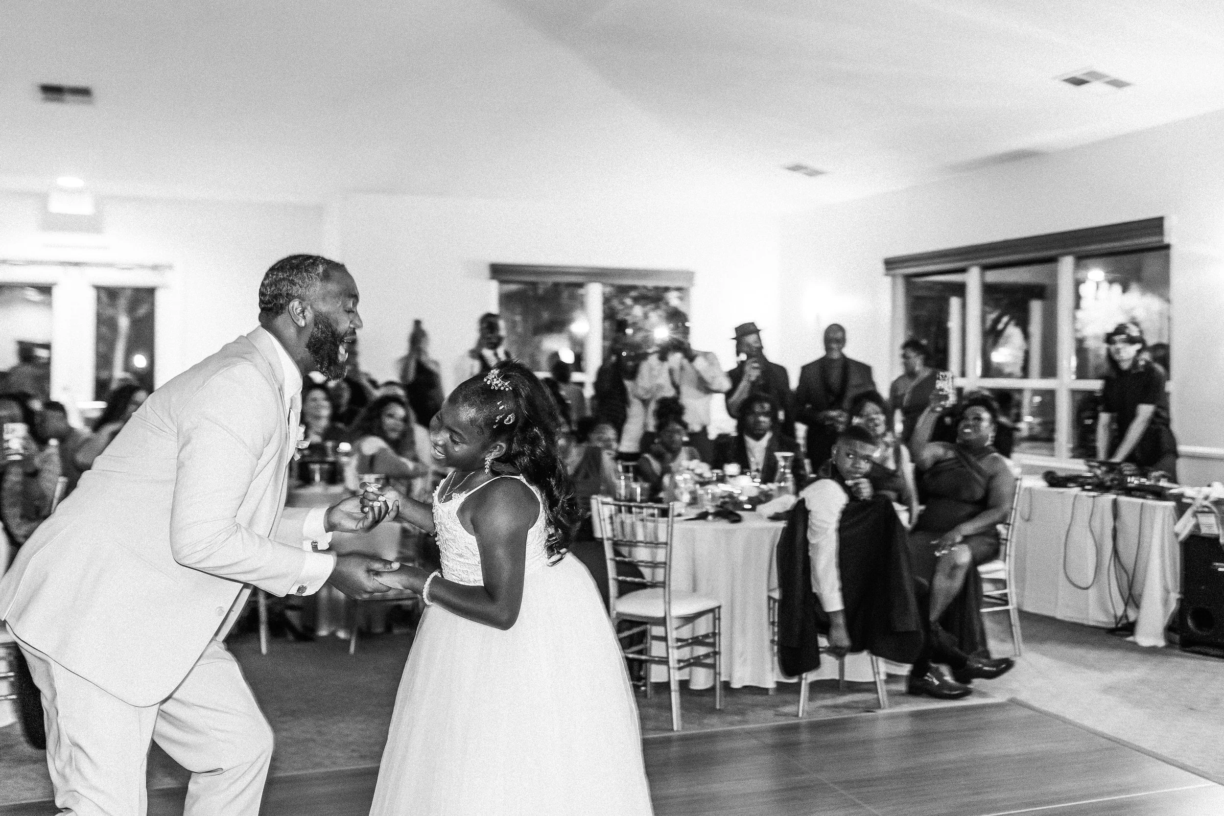 A man and a woman are dancing together at a wedding reception, surrounded by seated and standing guests in a decorated indoor venue.
