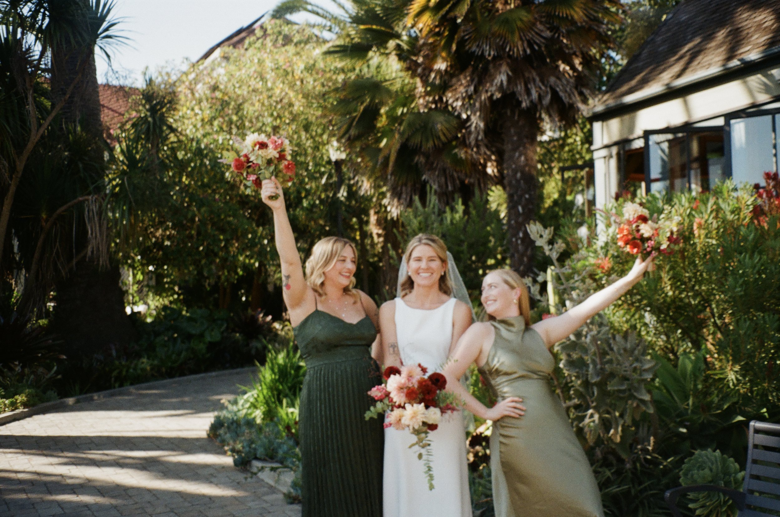 Three women in formal attire celebrating outdoors with bouquets of flowers, standing on a garden path surrounded by trees and plants.