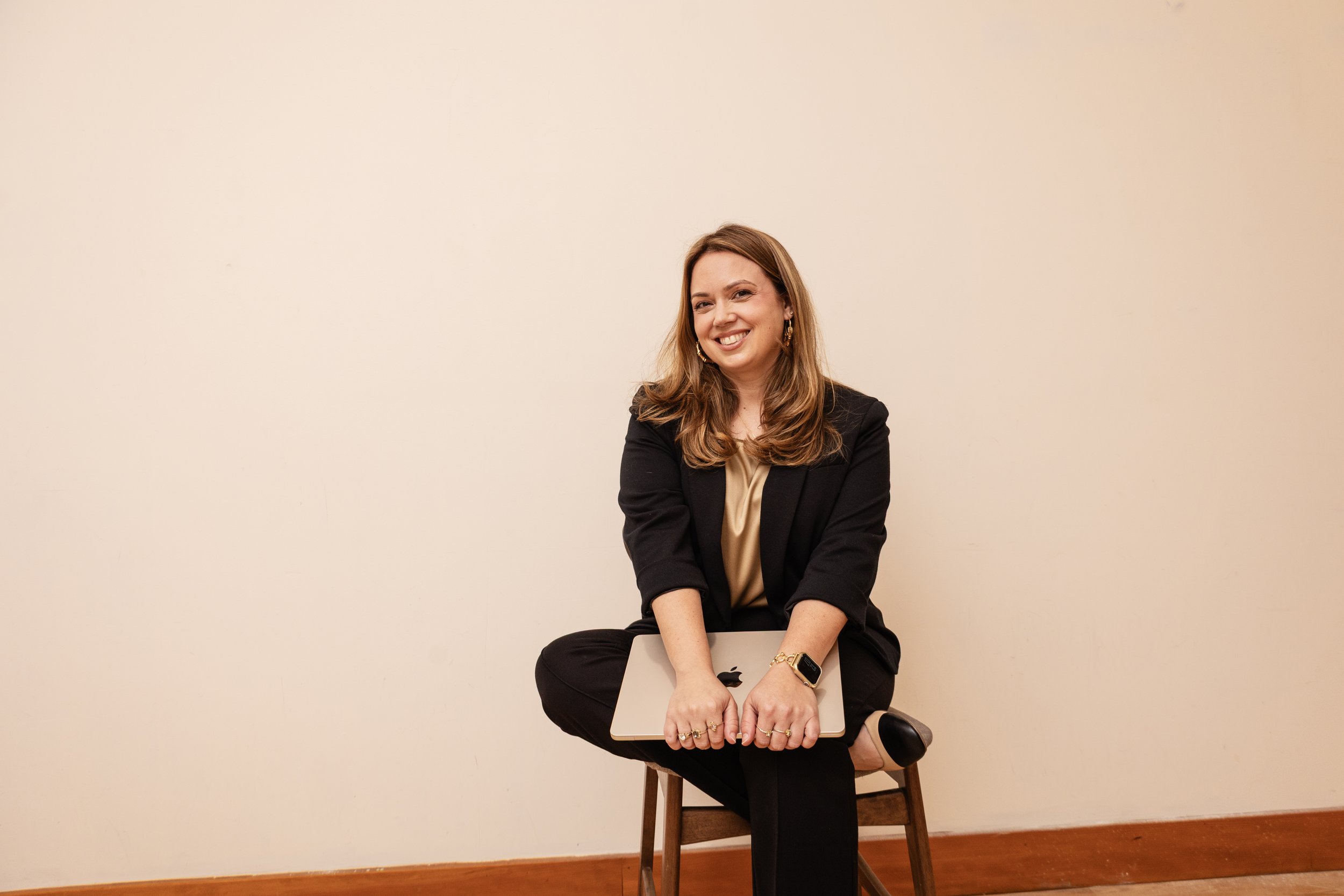 A woman with shoulder-length brown hair sitting on a stool in front of a plain beige wall. She is wearing a black blazer and pants, with a beige top underneath, and is smiling while holding a closed silver laptop on her lap. She has earrings, a watch