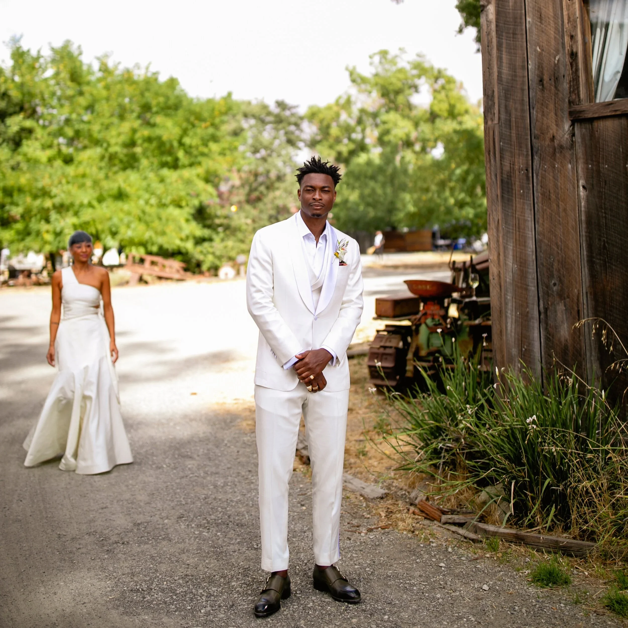 A man dressed in a white suit and black shoes standing outdoors during daytime, with a woman in a white dress behind him and trees in the background.