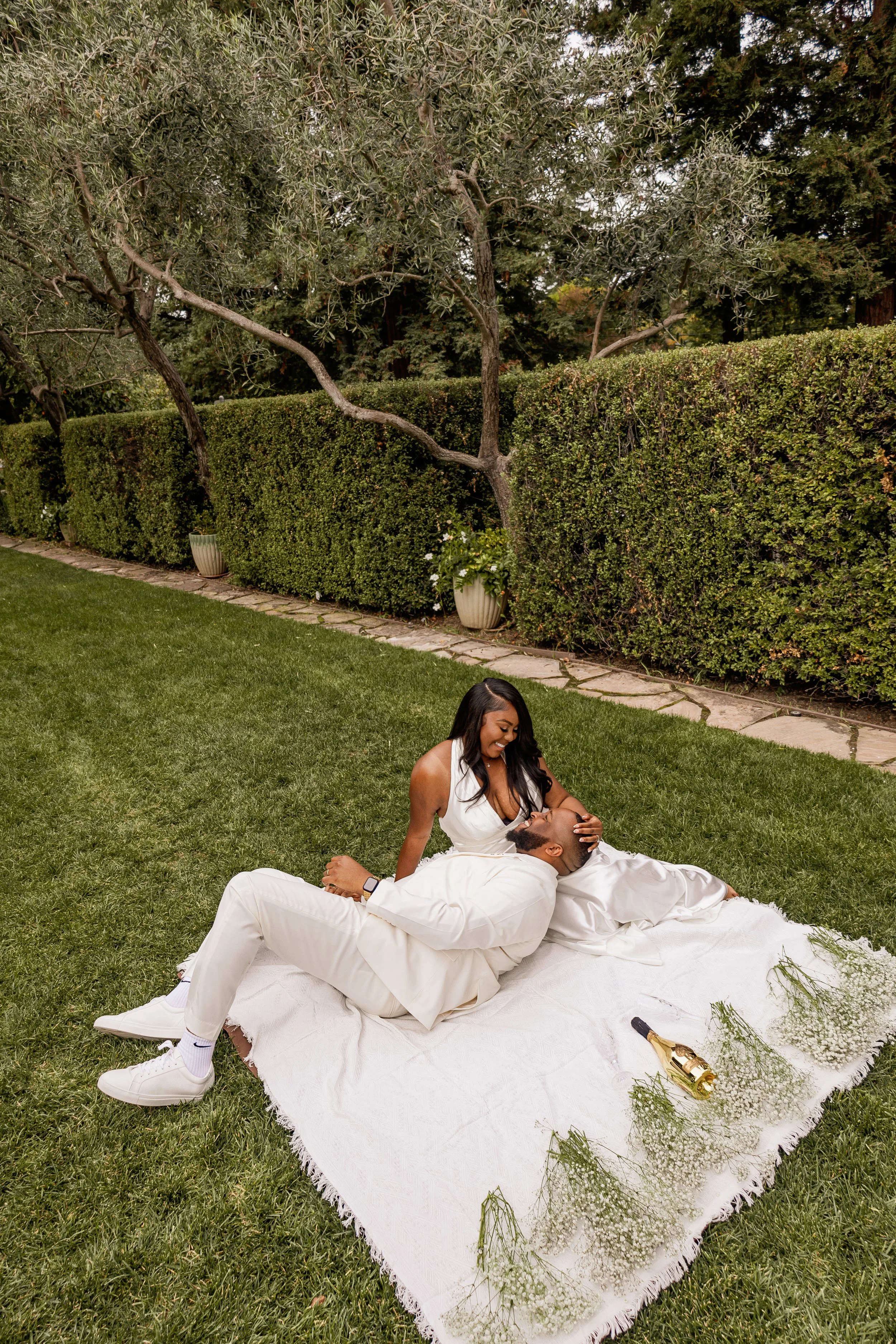 A woman and a man, both dressed in white, are sitting on a white blanket on the grass in a garden. The woman is smiling and leaning over the man, who is lying down, and she is holding his face. There are flowers and greenery around them, with a hedge and trees in the background. A champagne bottle wrapped in a gold foil and some greenery are on the blanket.