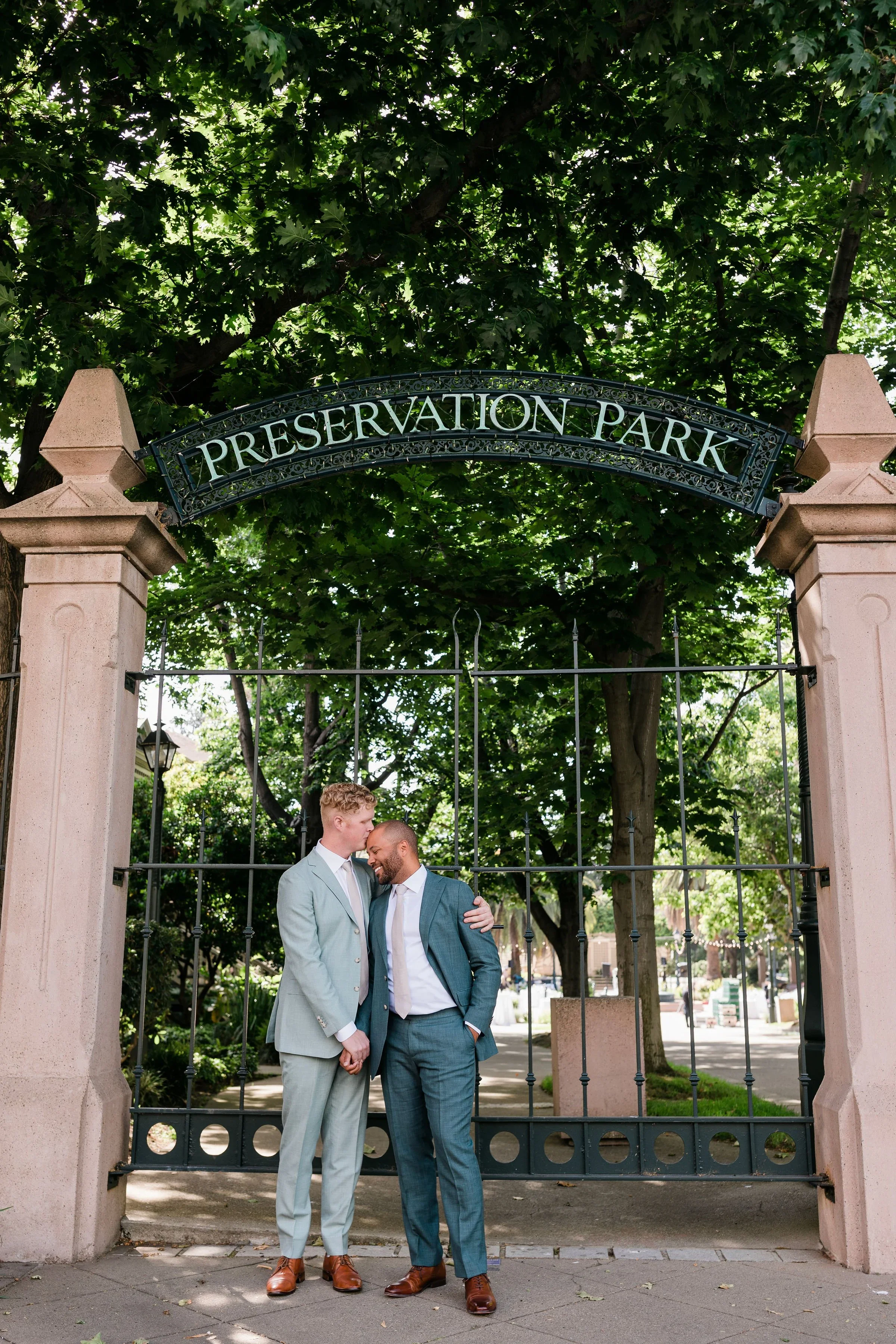 Two men in suits standing close together and smiling in front of a gate at Preservasion Park, with trees and a sidewalk visible in the background.