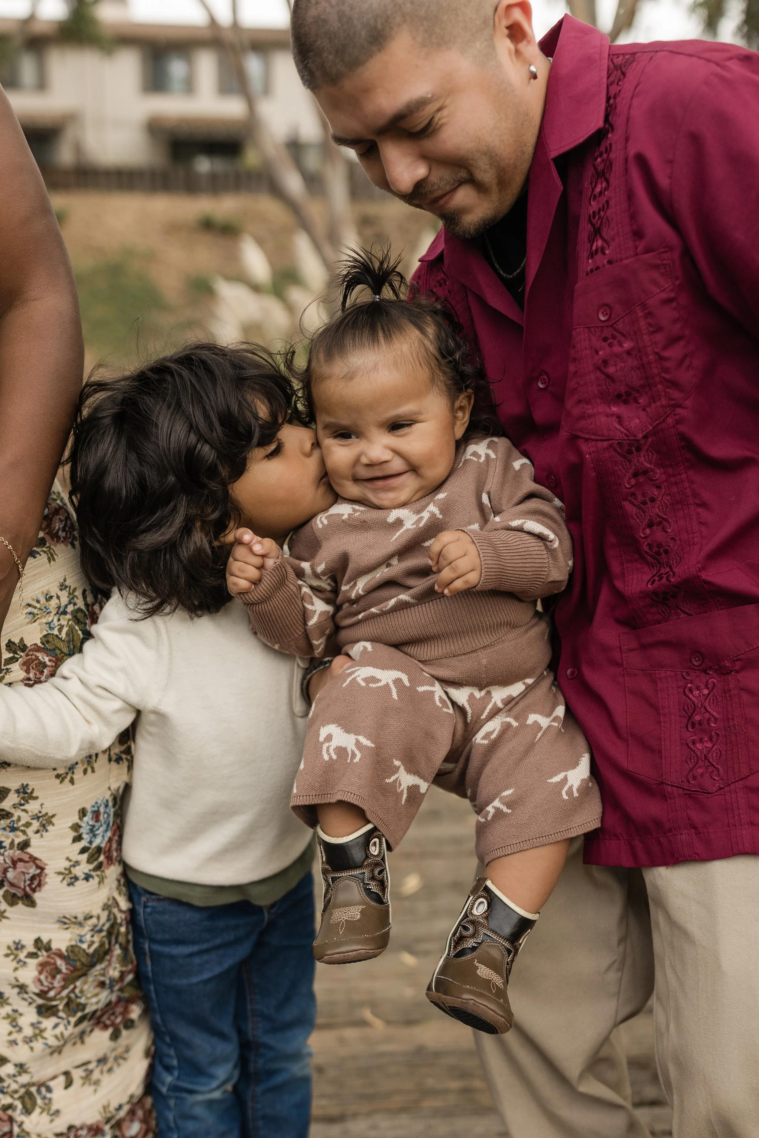 A man holding a smiling toddler girl, who is being kissed on her cheek by a woman. Two children are visible, with one girl and one boy, on a wooden deck outdoors.
