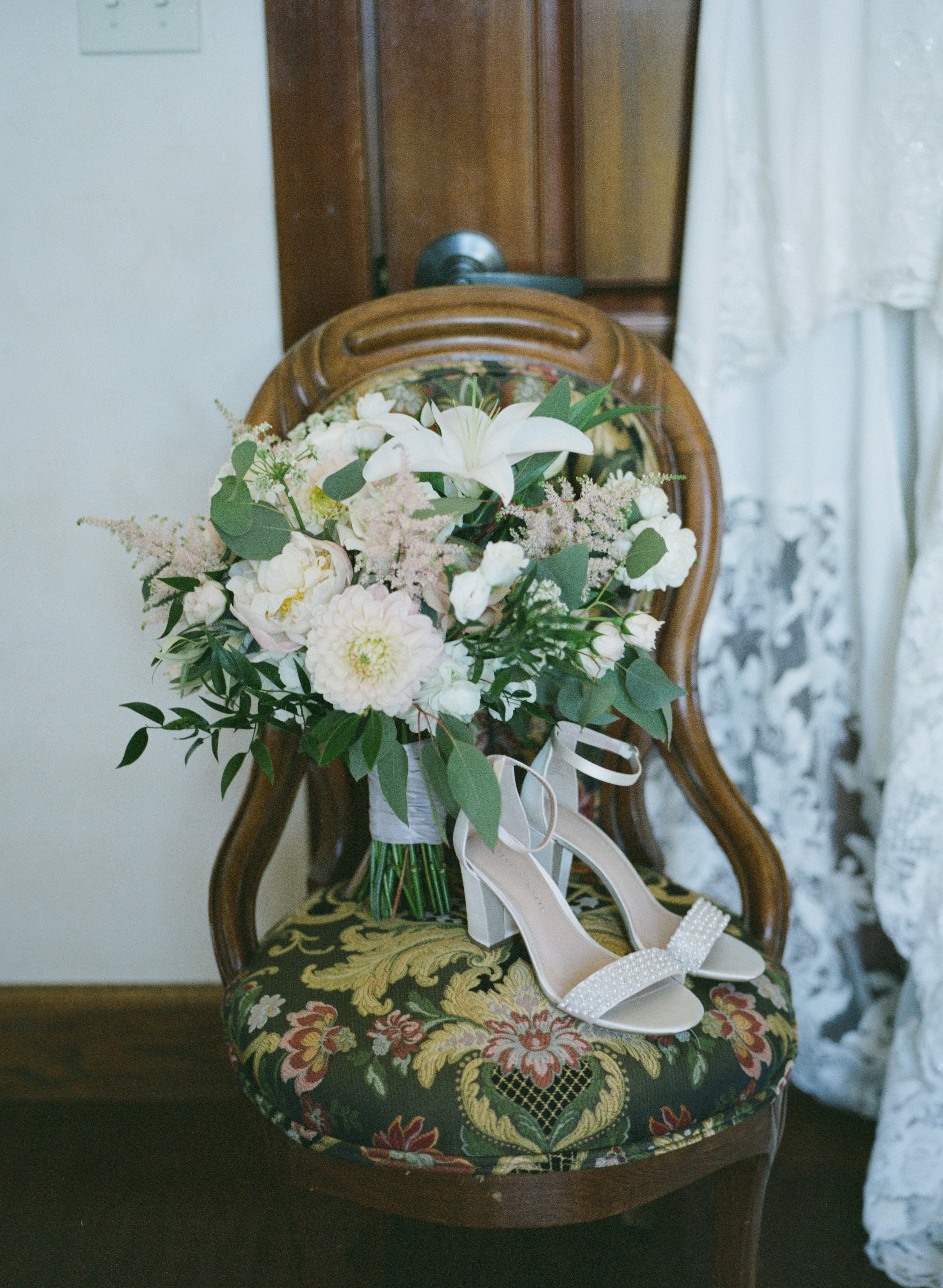 A bouquet of white and pastel-colored flowers resting on a vintage wooden chair with a floral-patterned cushion. A pair of white high-heeled shoes with pearl embellishments are placed on the chair next to the bouquet.