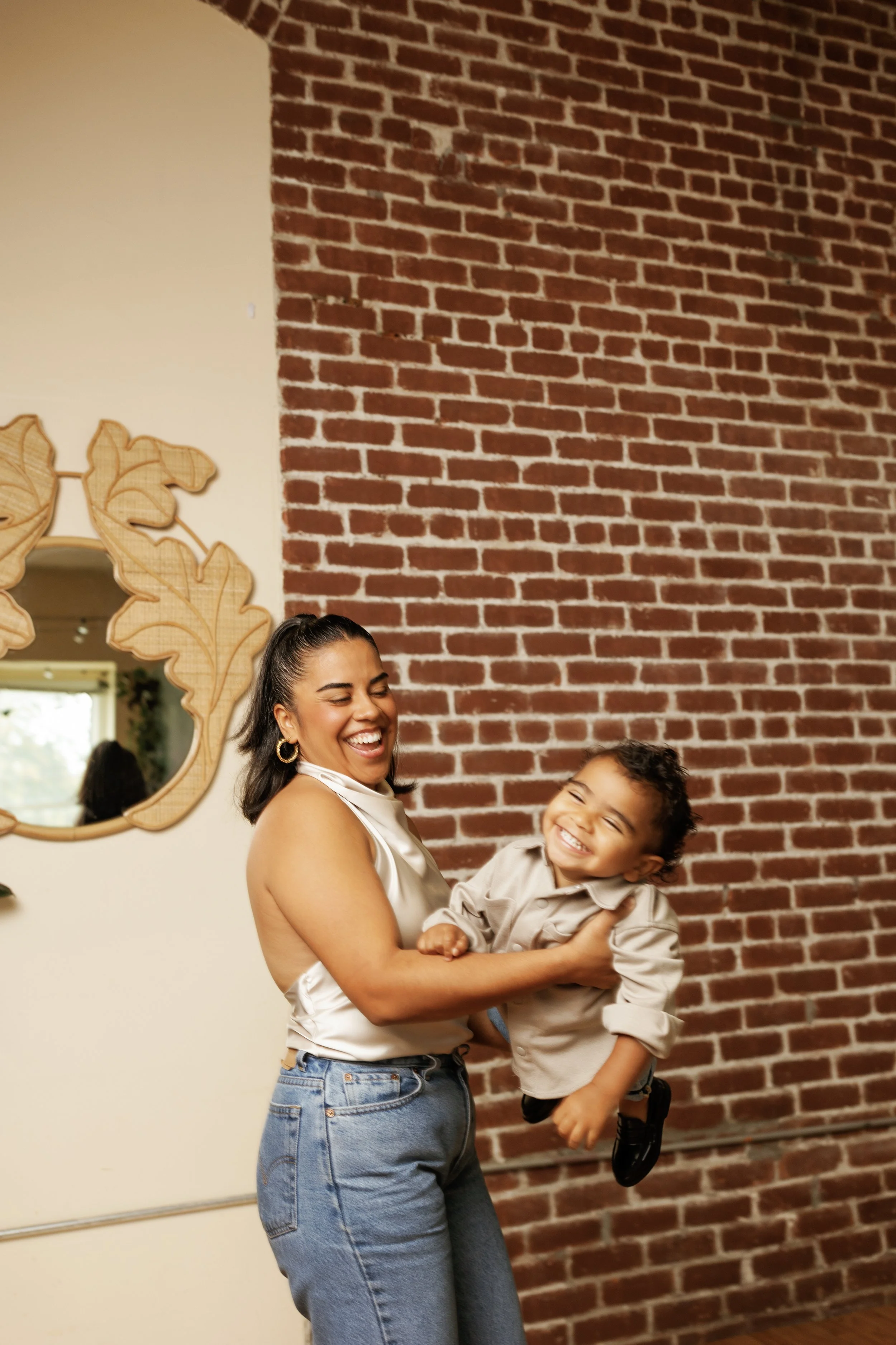 A woman and a young boy smiling and laughing as she holds him, standing in front of a brick wall and a mirror decorated with wooden leaves.
