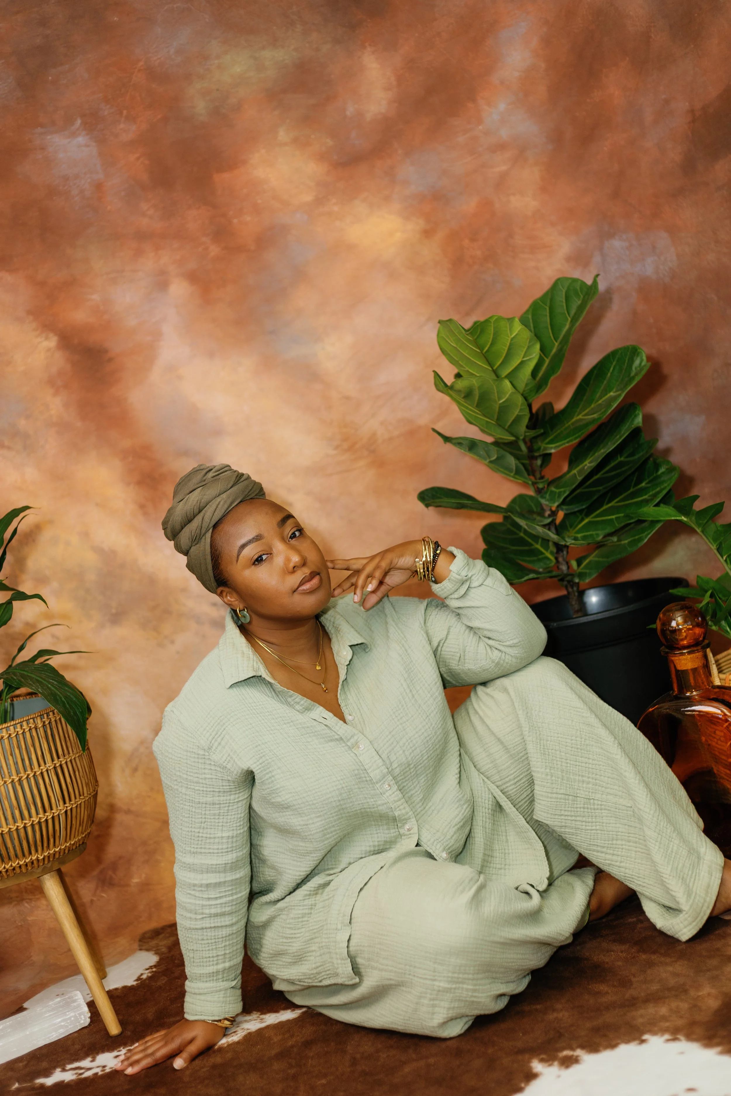 A woman with dark skin, wearing a light green, textured outfit and jewelry, is sitting on the floor next to a large plant with broad, green leaves. She is posed with one hand near her face and the other hand on the floor, against a warm, textured bro