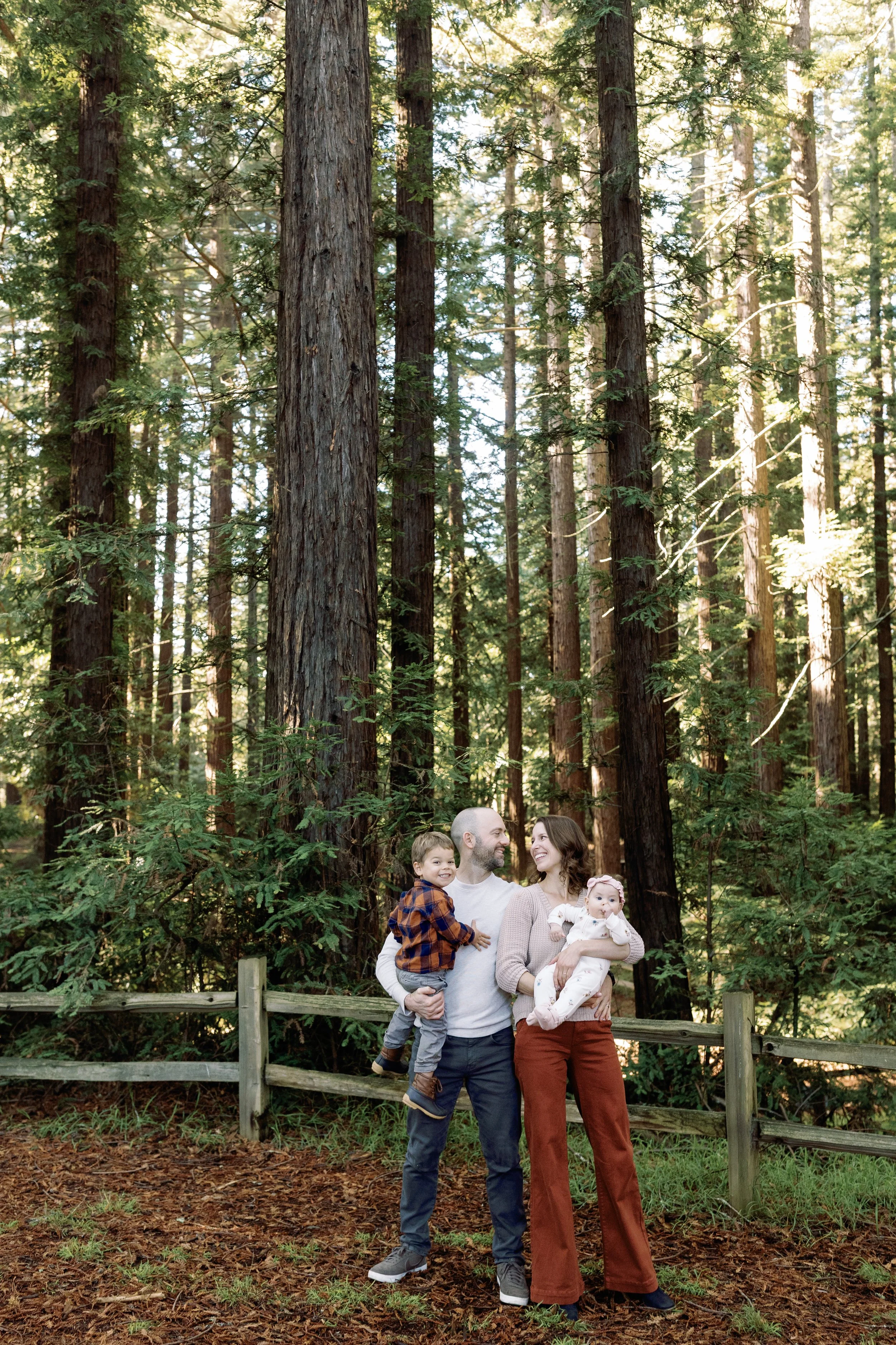 A family of four standing outdoors in a forest near a wooden fence, with tall trees and sunlight filtering through the leaves behind them. The father is holding a young boy, and the mother is holding a baby girl, all smiling and looking happy.