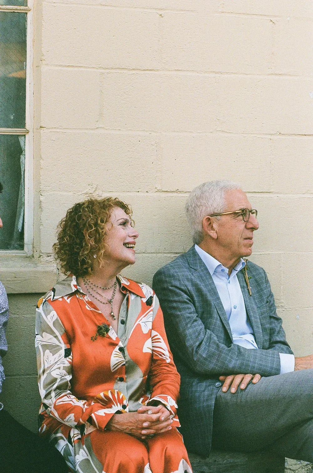 A woman with curly red hair in an orange, white, and black patterned blouse smiling and sitting next to a man with light gray hair and glasses, wearing a blue checkered blazer and white shirt, sitting with arms crossed against a beige wall.