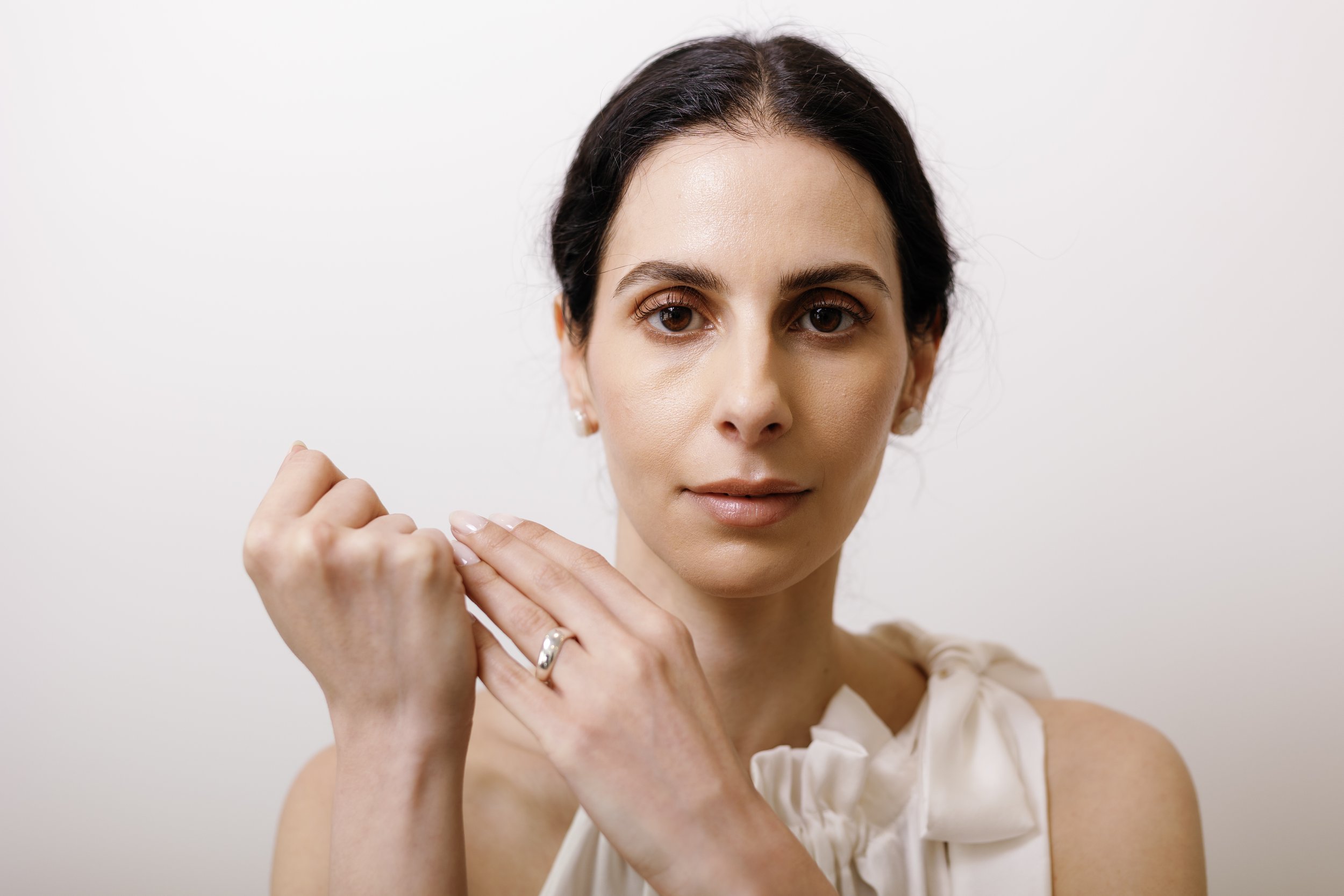 A close-up of a woman with dark hair, brown eyes, and light skin, holding a small object near her face with both hands.