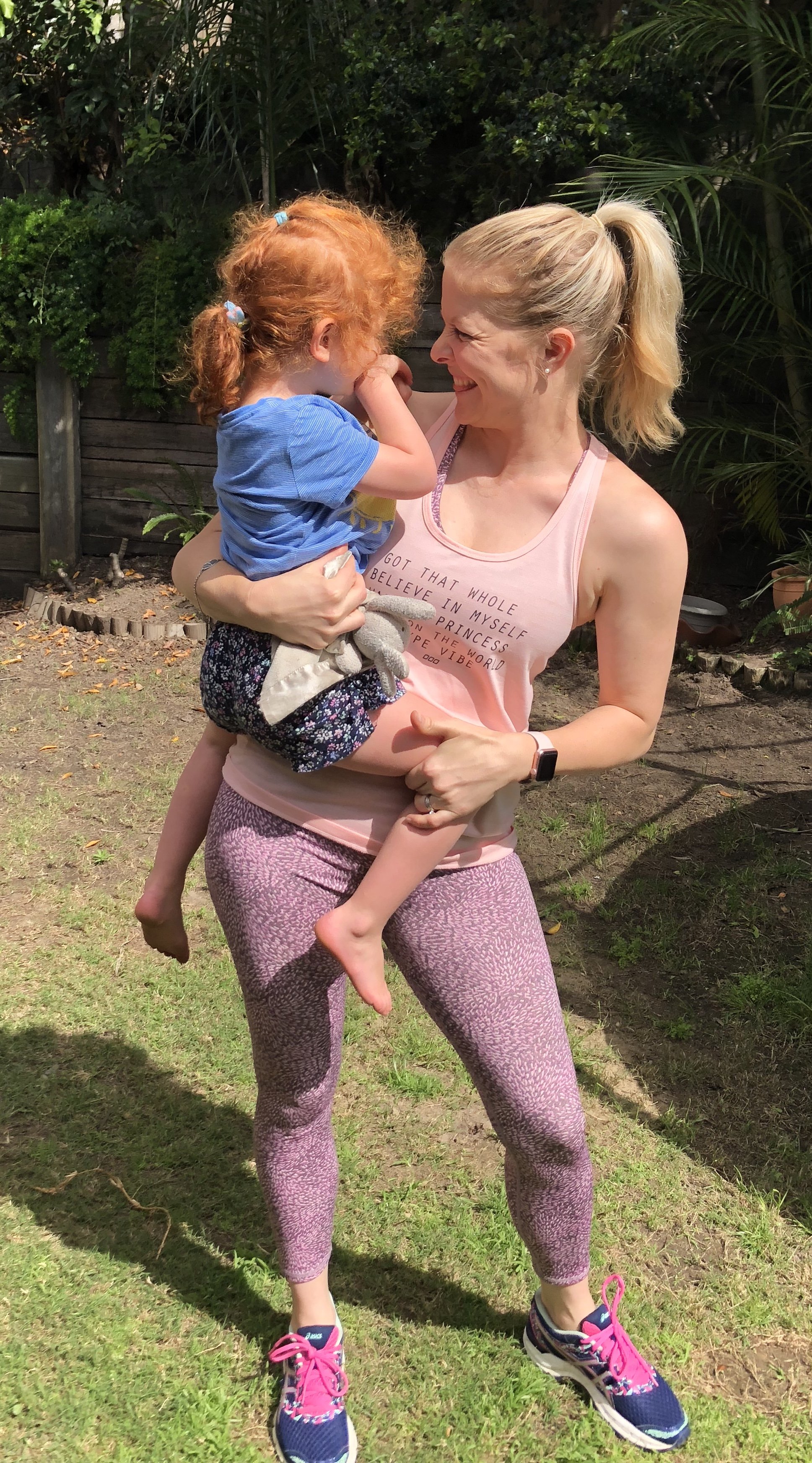 A woman with blonde hair in a ponytail holding a young red-haired girl in a backyard garden. Both are smiling and laughing, wearing casual workout clothes, with greenery in the background.