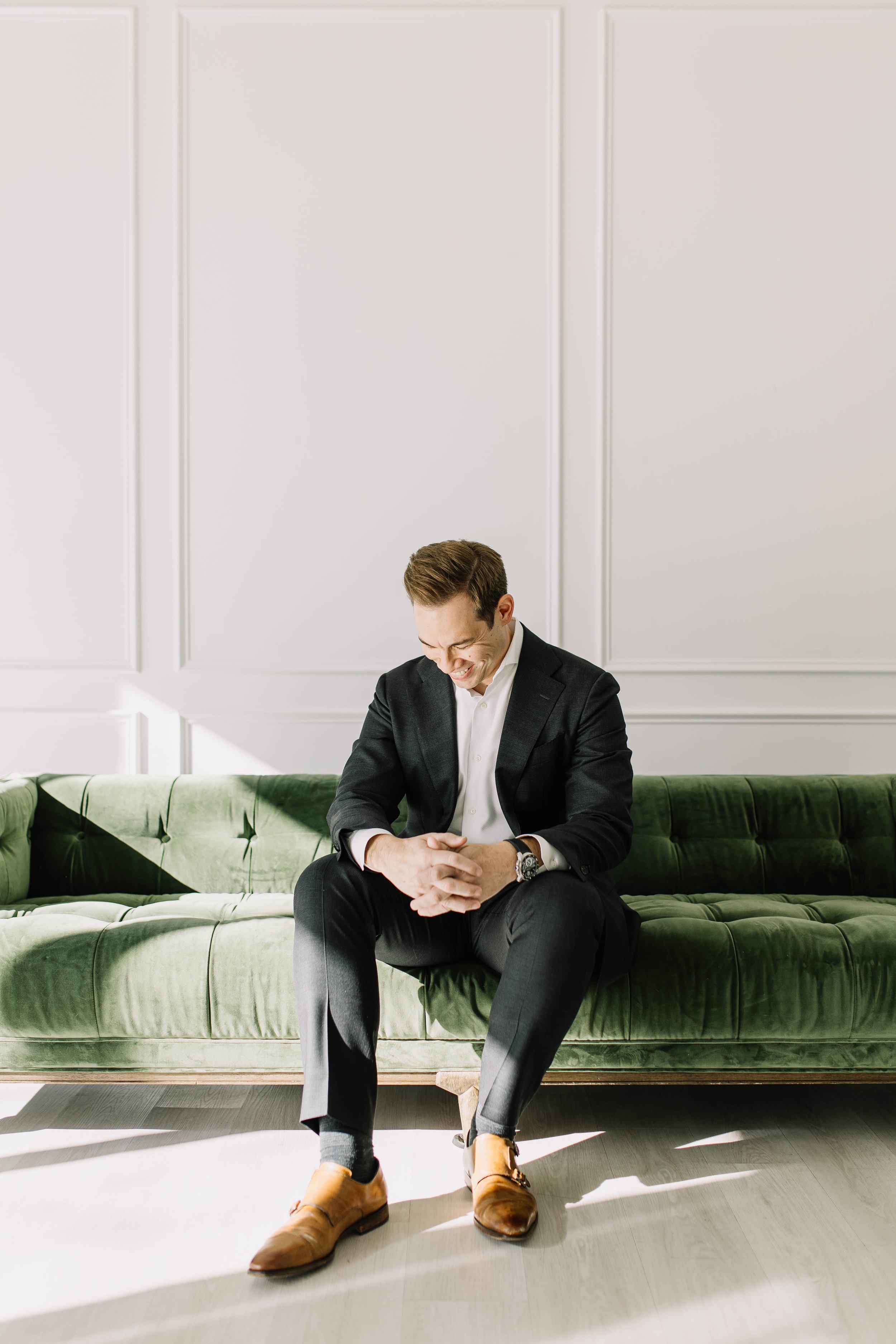 A young man in a black suit and brown dress shoes sitting on a green velvet couch, laughing with his head bowed and hands clasped on his lap, in a room with white paneled walls.