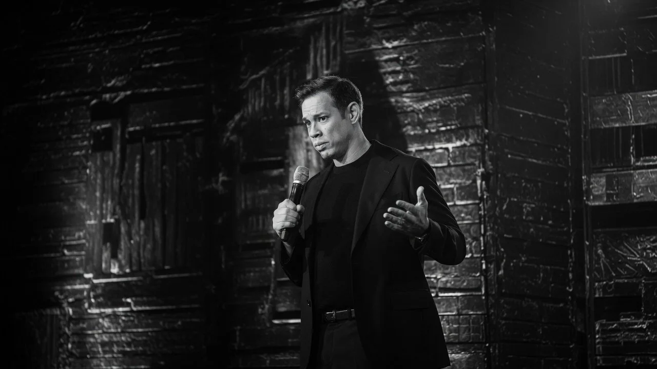 A man holding a microphone performing on stage in a black blazer and shirt, with a dark wooden textured wall behind him.
