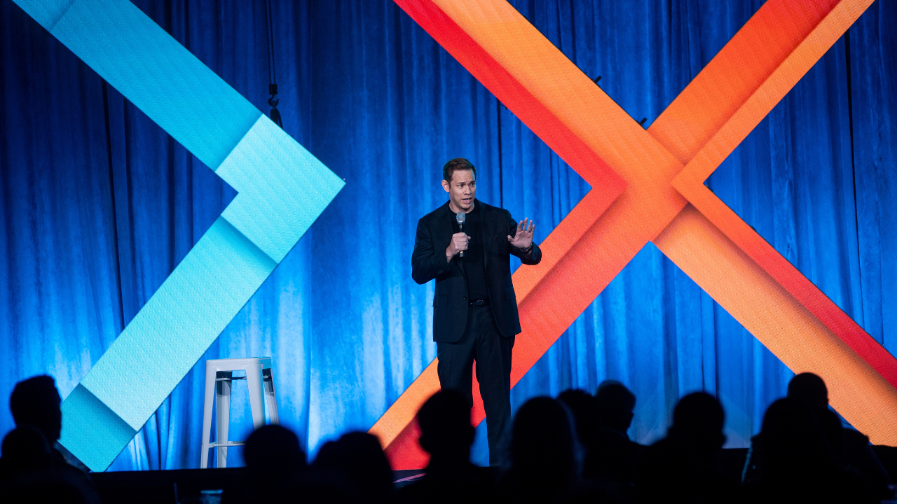 A man in a suit giving a presentation on stage with blue curtains and colorful geometric decorations, audience silhouetted in front.