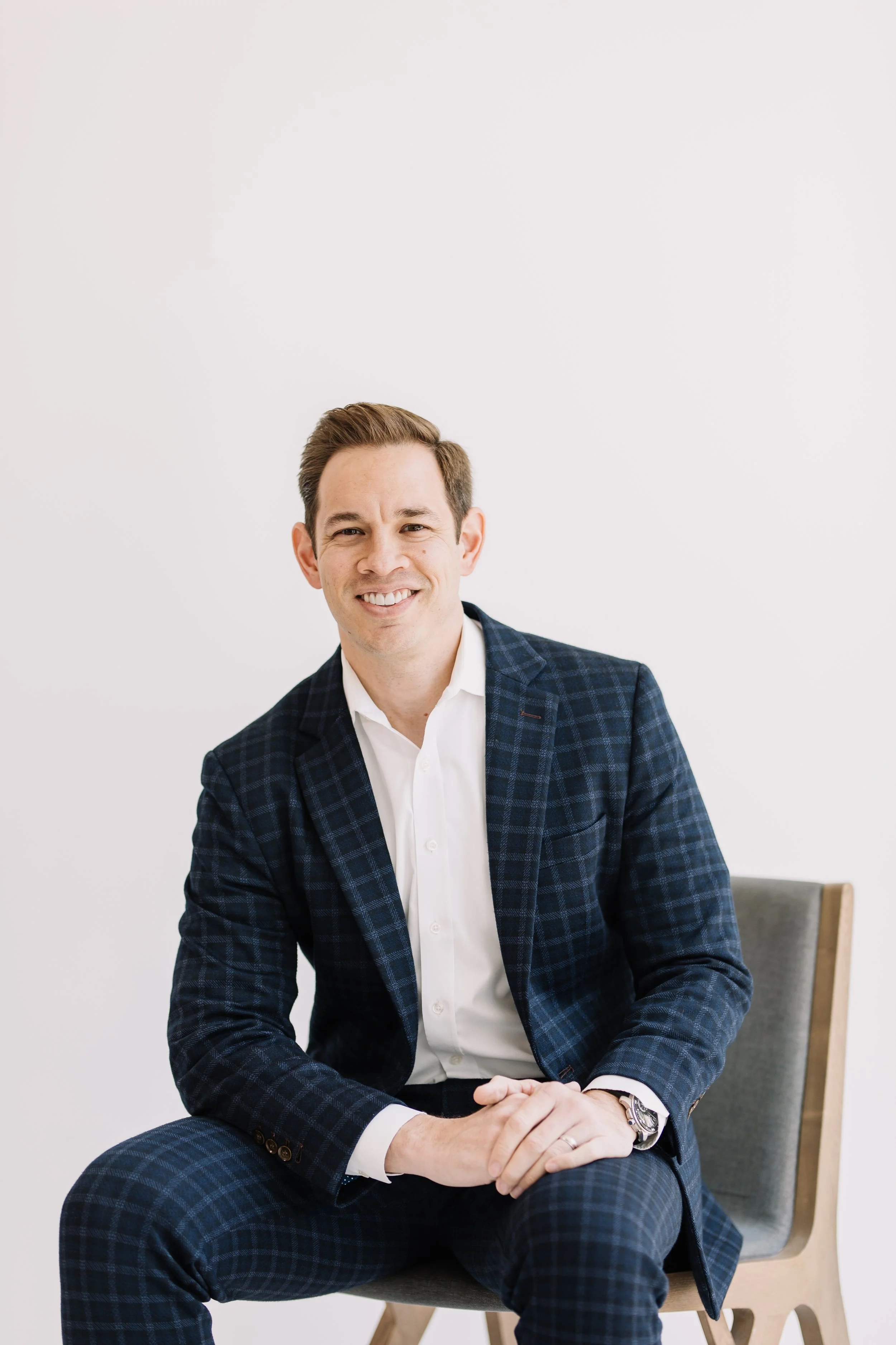 A smiling man in a navy blue checkered suit and white shirt sits on a gray chair against a plain white wall.