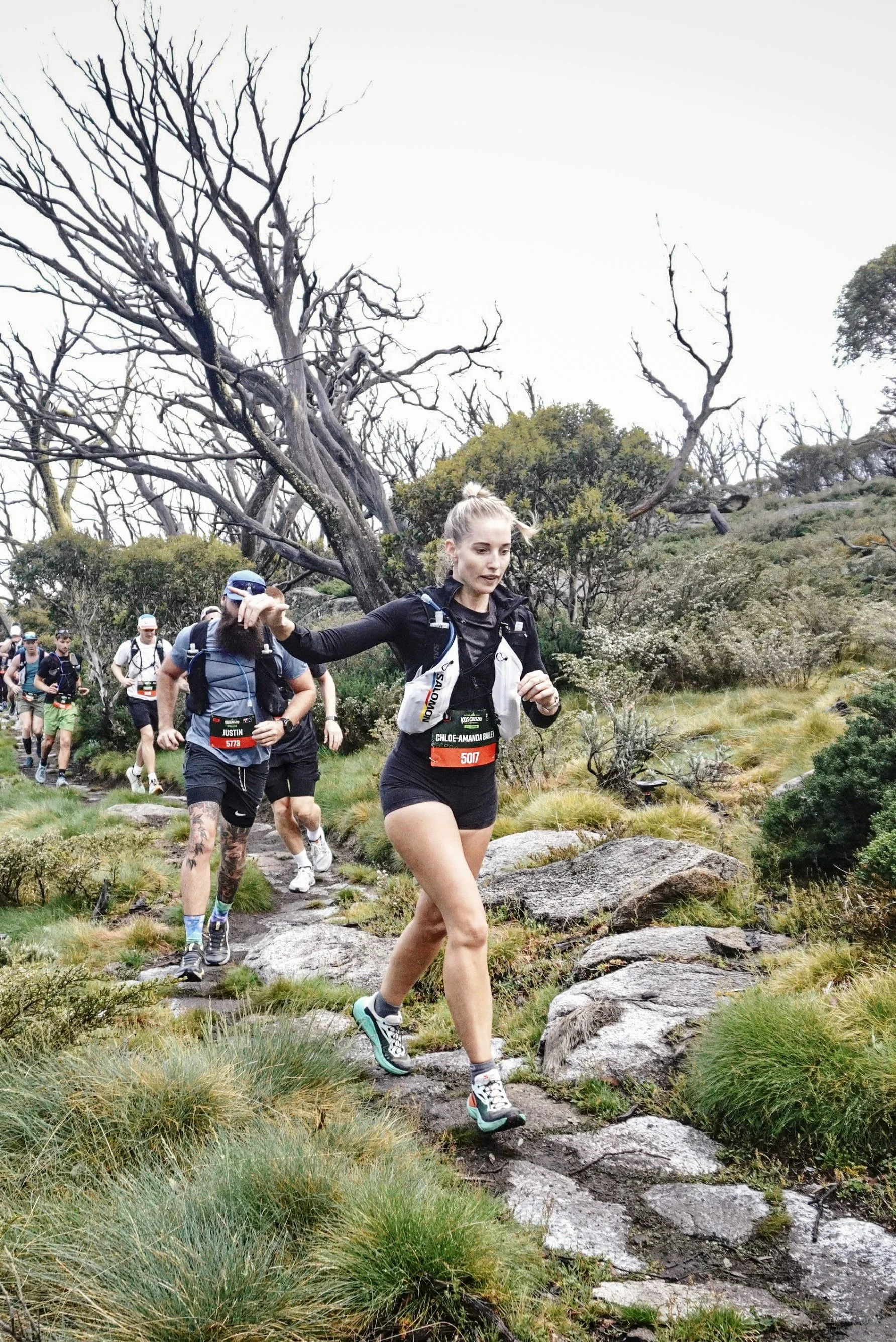 A group of trail runners, including a woman in black athletic wear, running on a rocky trail in a forested area with leafless trees and green bushes.