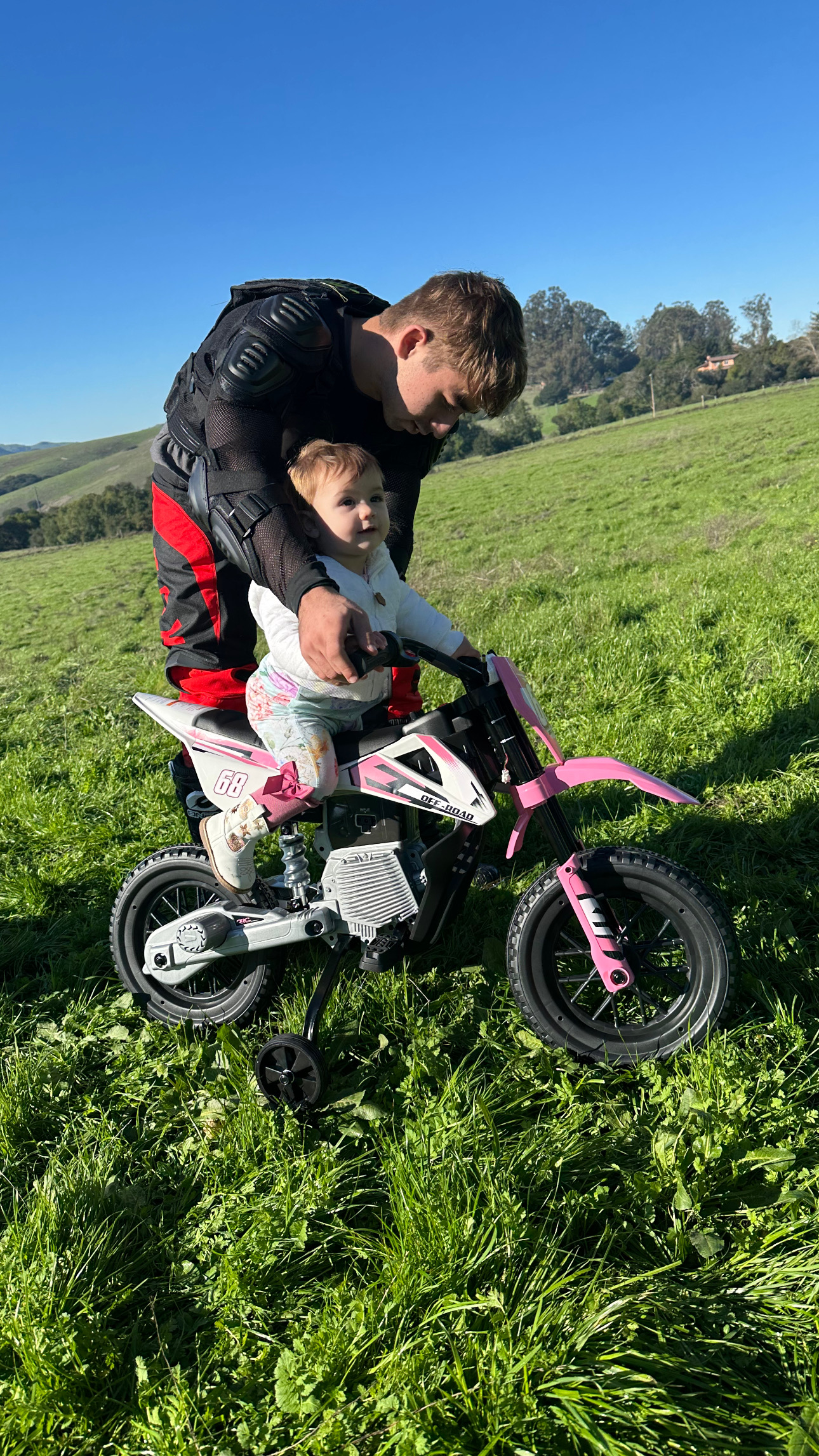 A man helping a young girl learn to ride a pink and white kid's bicycle with training wheels in a grassy field on a sunny day.