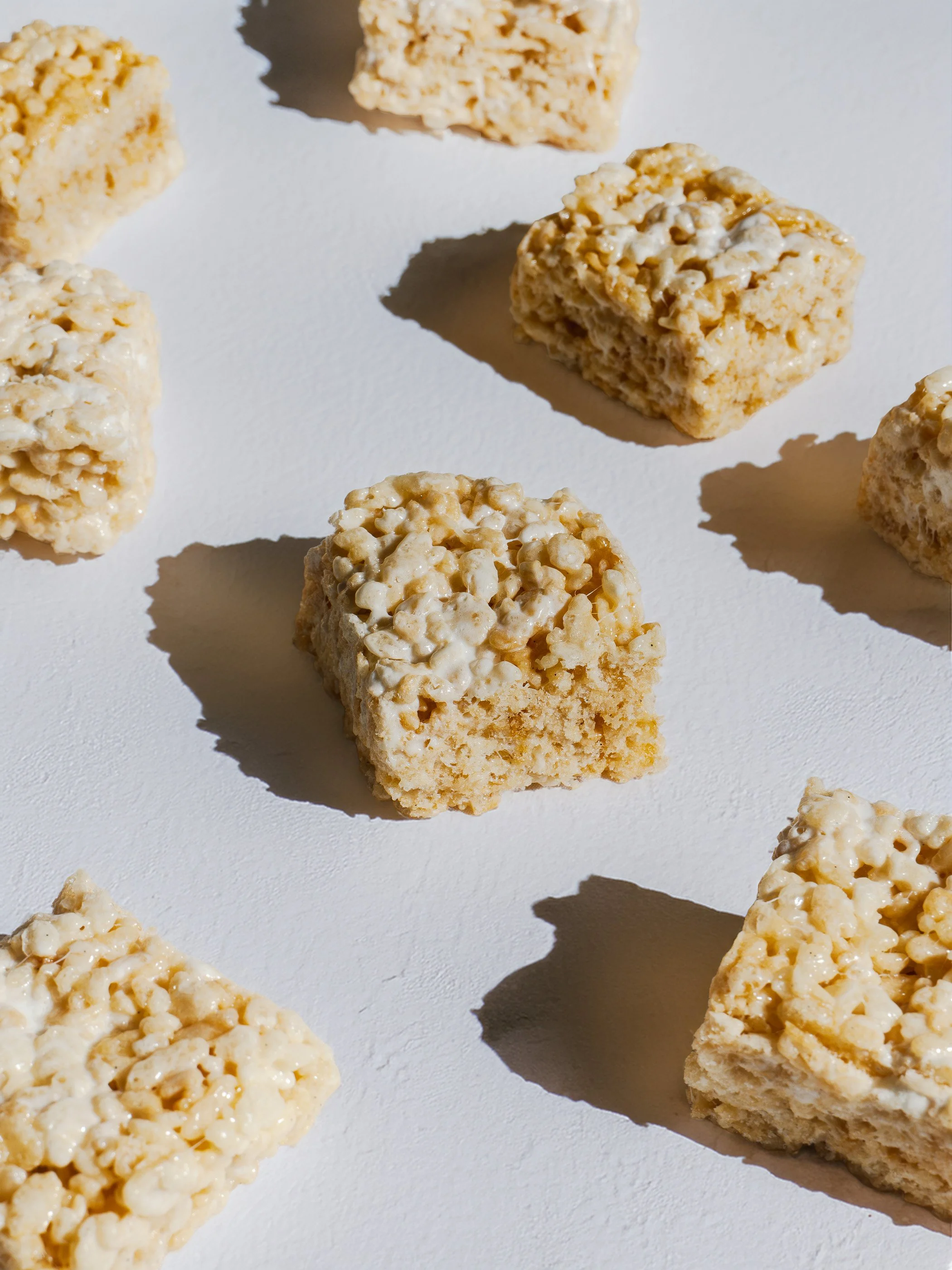 Several rice cereal treats with marshmallows are arranged on a white surface, casting shadows in bright sunlight.