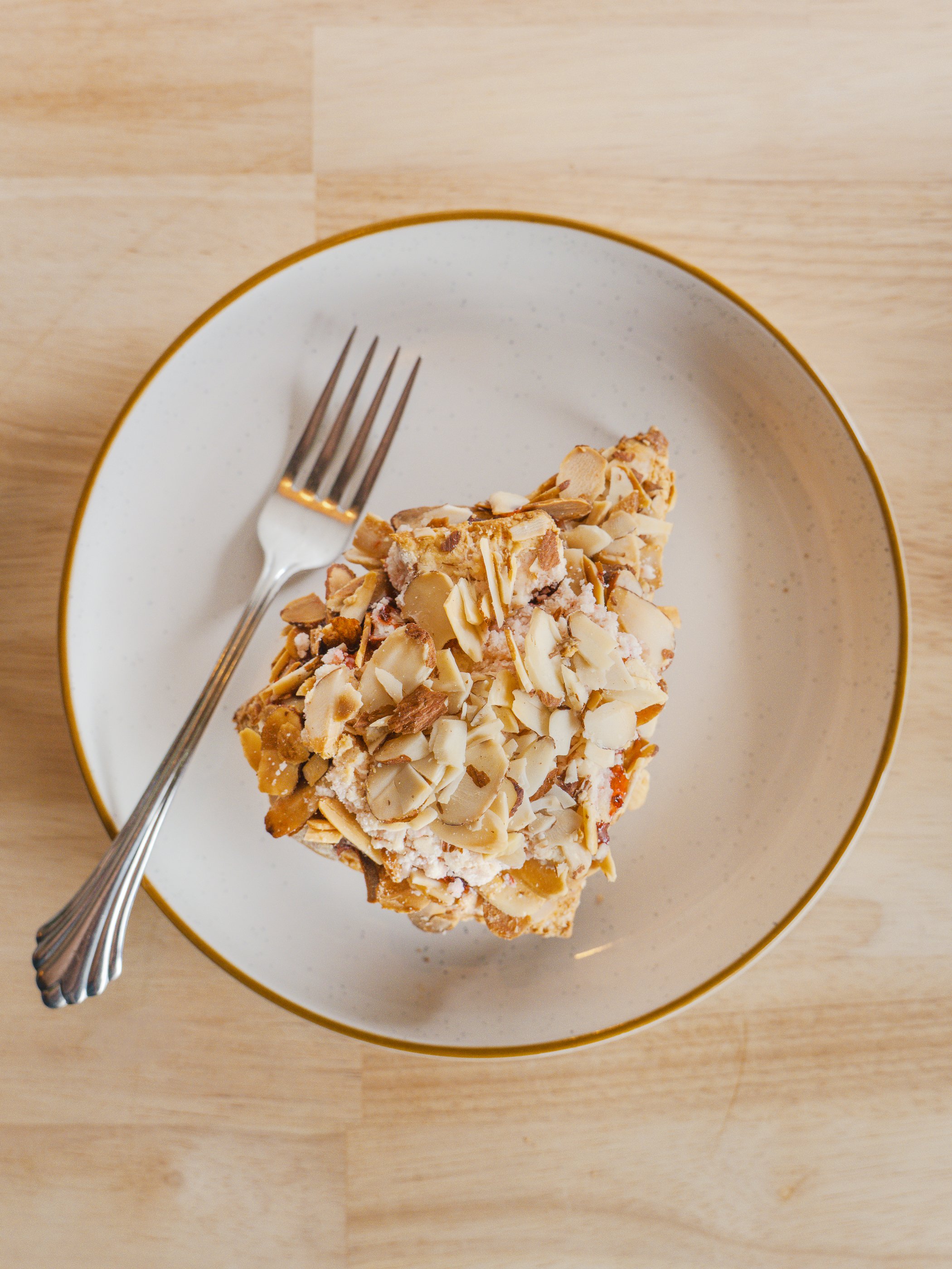 A slice of almond cake on a white dish with gold trim, placed on a wooden table, with a fork beside it.