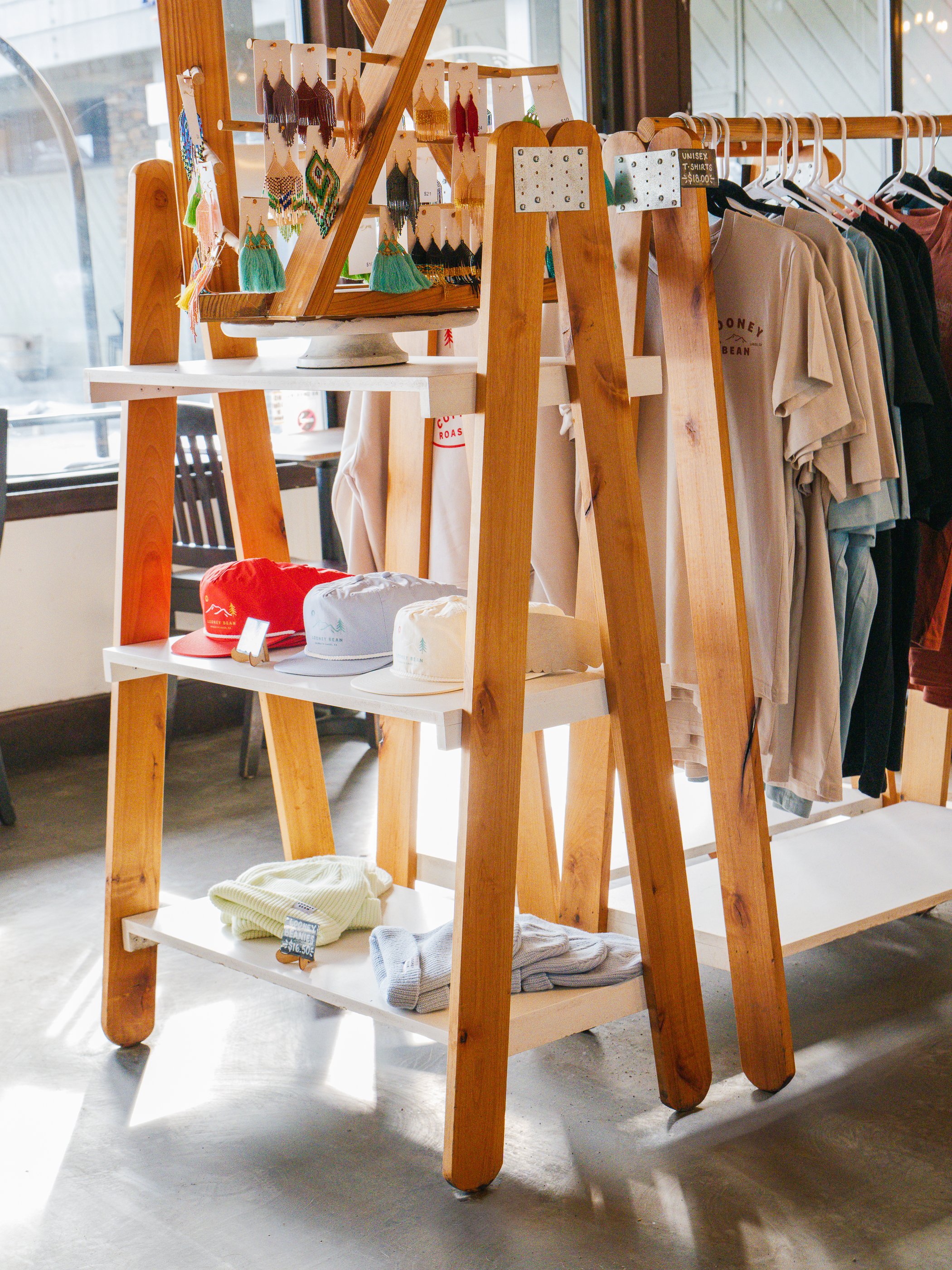 Display shelf in a retail clothing store with hats, beaded earrings, and folded sweaters, next to hanging T-shirts near a window.