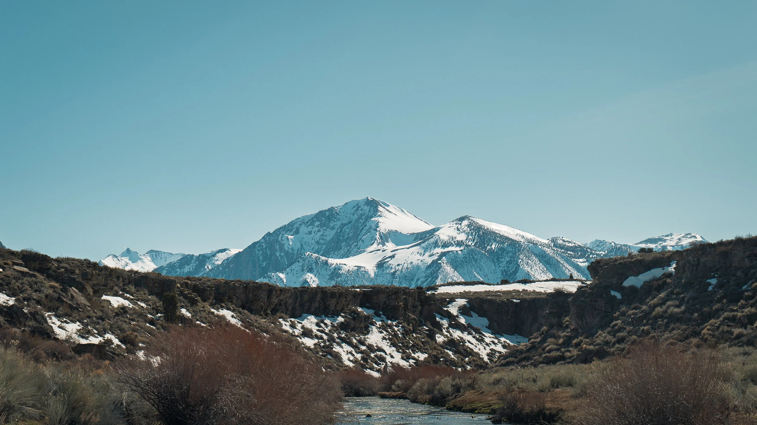 Snow-capped mountains above a river flowing through a valley with sparse shrubs and bushes.
