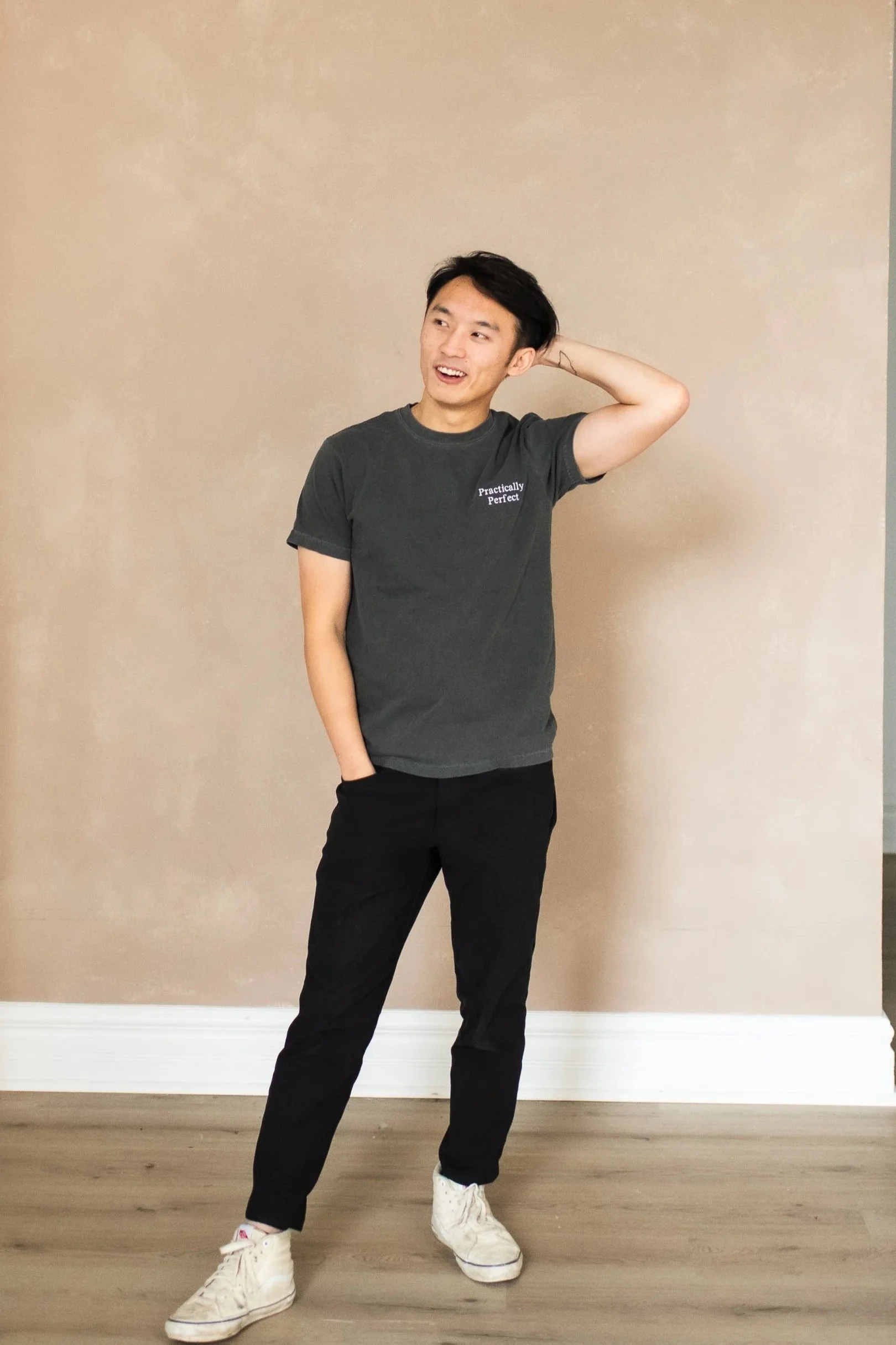 A young man with black hair wearing a dark gray T-shirt with white text and black pants, standing in front of a beige wall, smiling, with one hand behind his head and the other in his pocket.