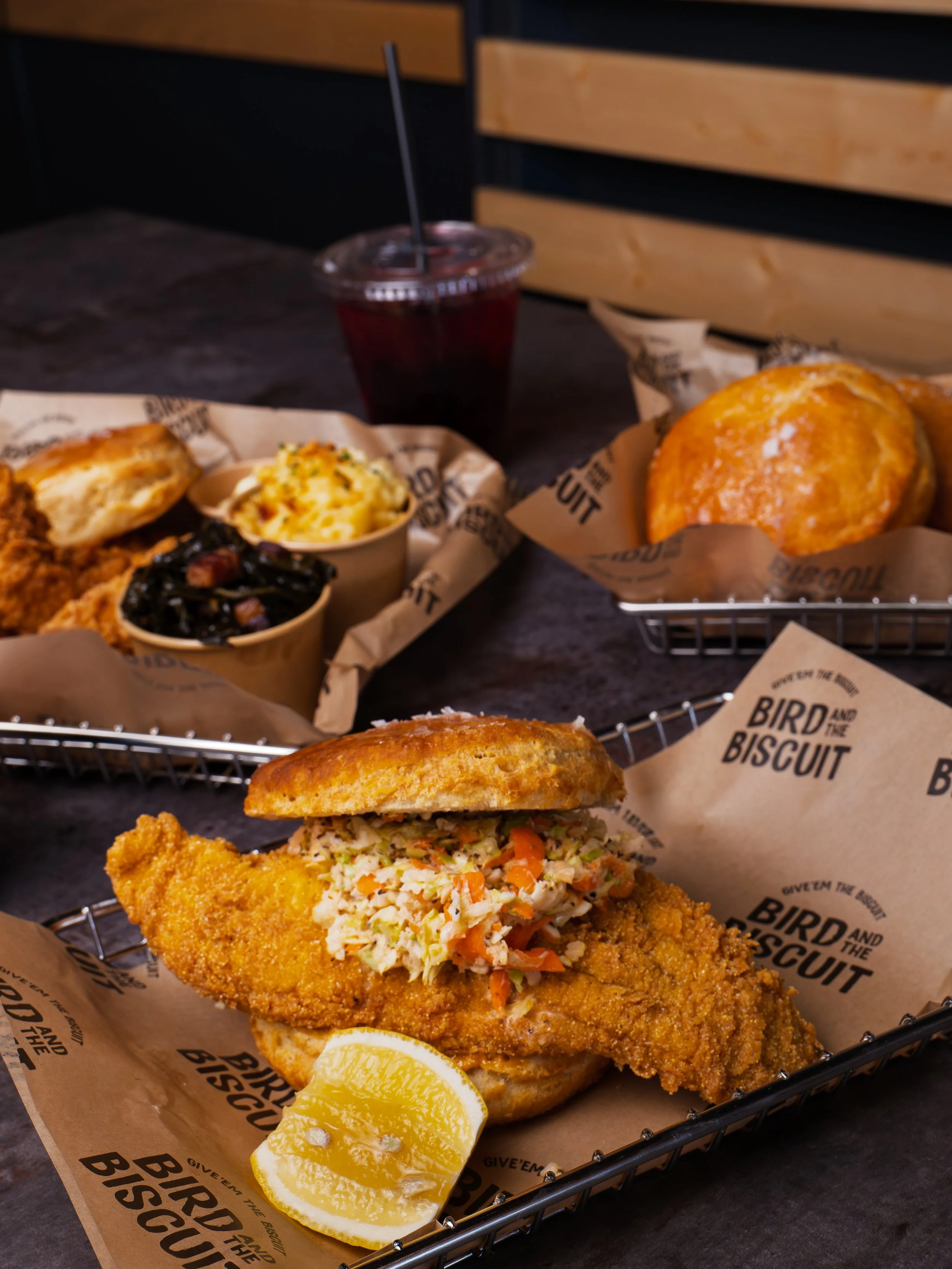A fried fish sandwich with coleslaw and a lemon wedge on a basket, with a side of fried chicken, mashed potatoes with gravy, a biscuit, and a soda in the background on a dark wood table.