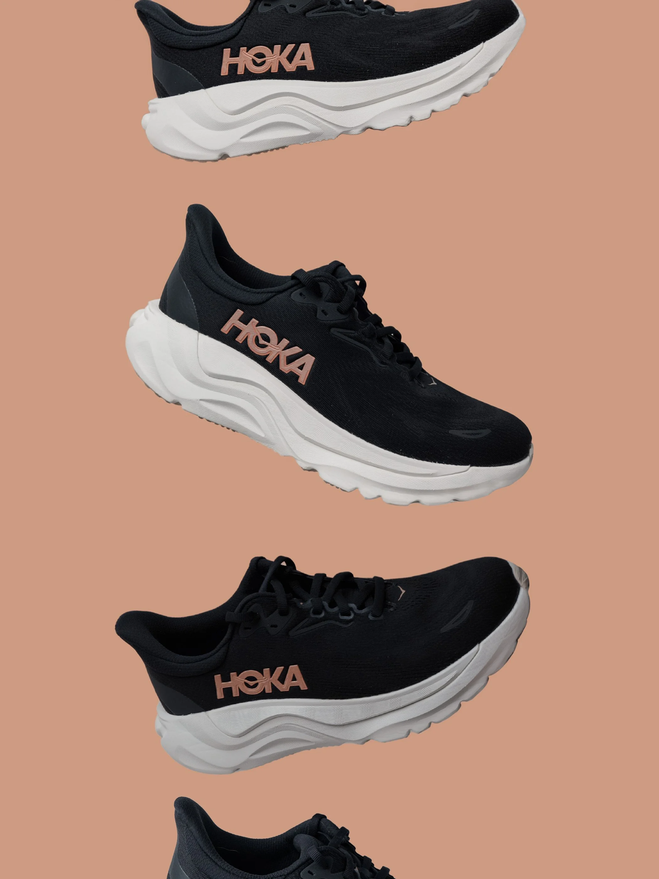 Stacked black running shoes with white soles and pink HOKA logos on a peach background.