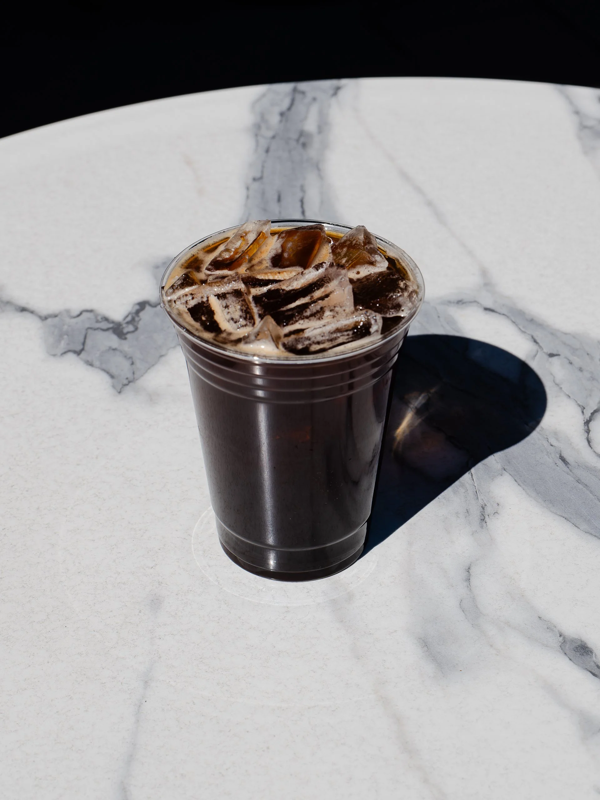 A plastic cup of iced coffee with ice cubes on a white marble table.