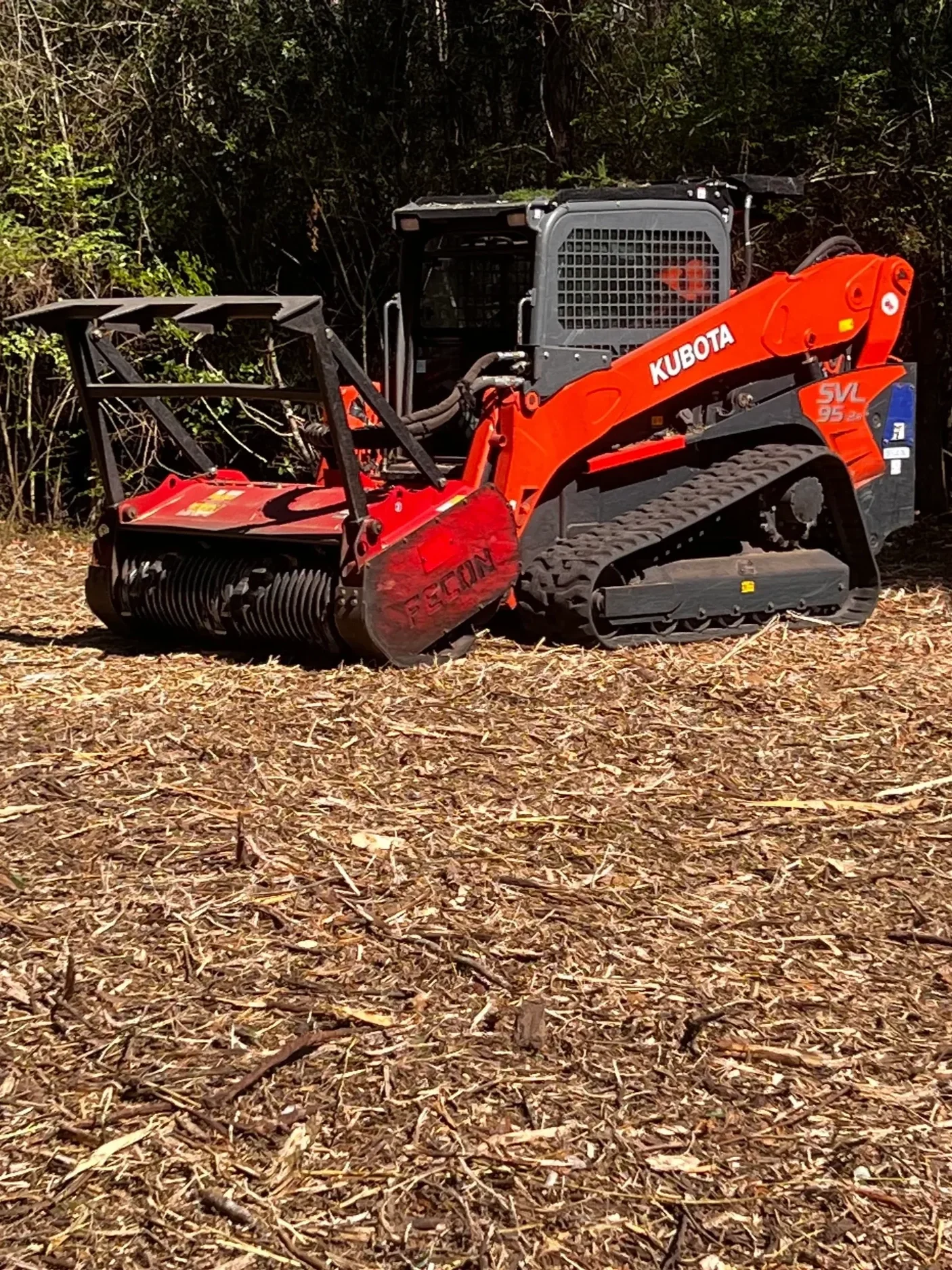 A Kubota SVL 95 track loader on dry, leaf-covered ground with bushes in the background.
