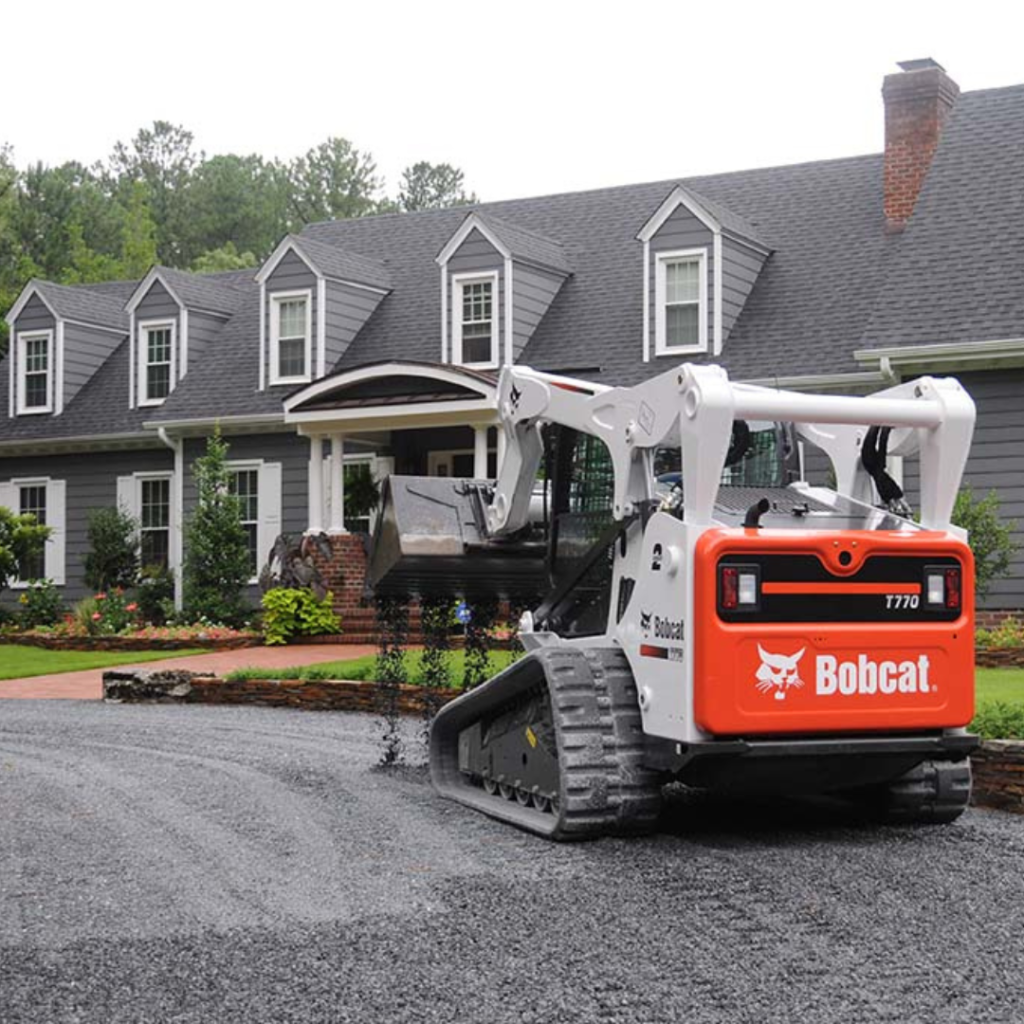 A Bobcat T770 compact track loader with black tracks and orange and white body, leveling a gravel driveway in front of a large gray house with dormer windows and a brick chimney.