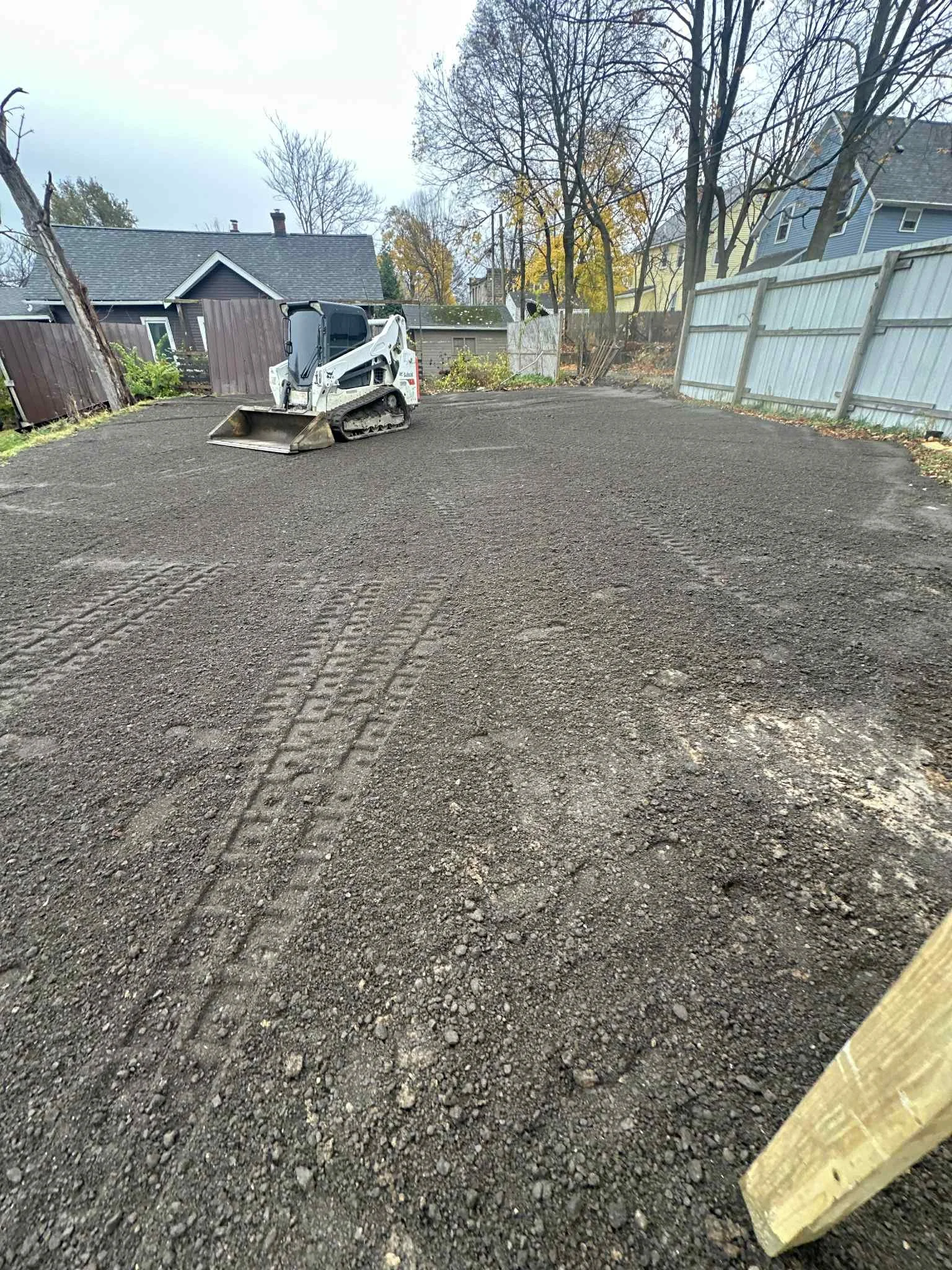 A gravelly backyard with a small Bobcat skid-steer loader, fenced in with wooden fences and trees with fall foliage.