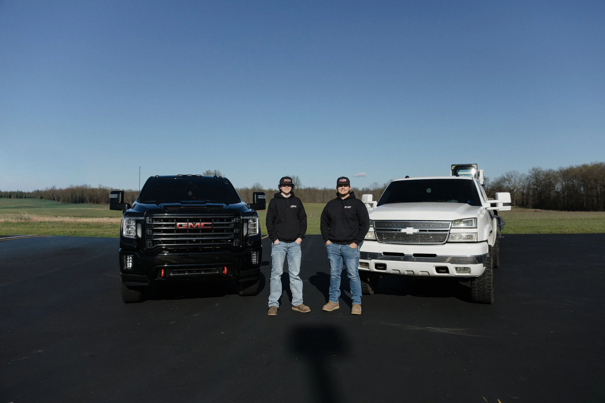 Two men in black hoodies and jeans standing outdoors between a black GMC truck and a white Chevrolet truck on a paved area with field and trees in the background under a clear blue sky.
