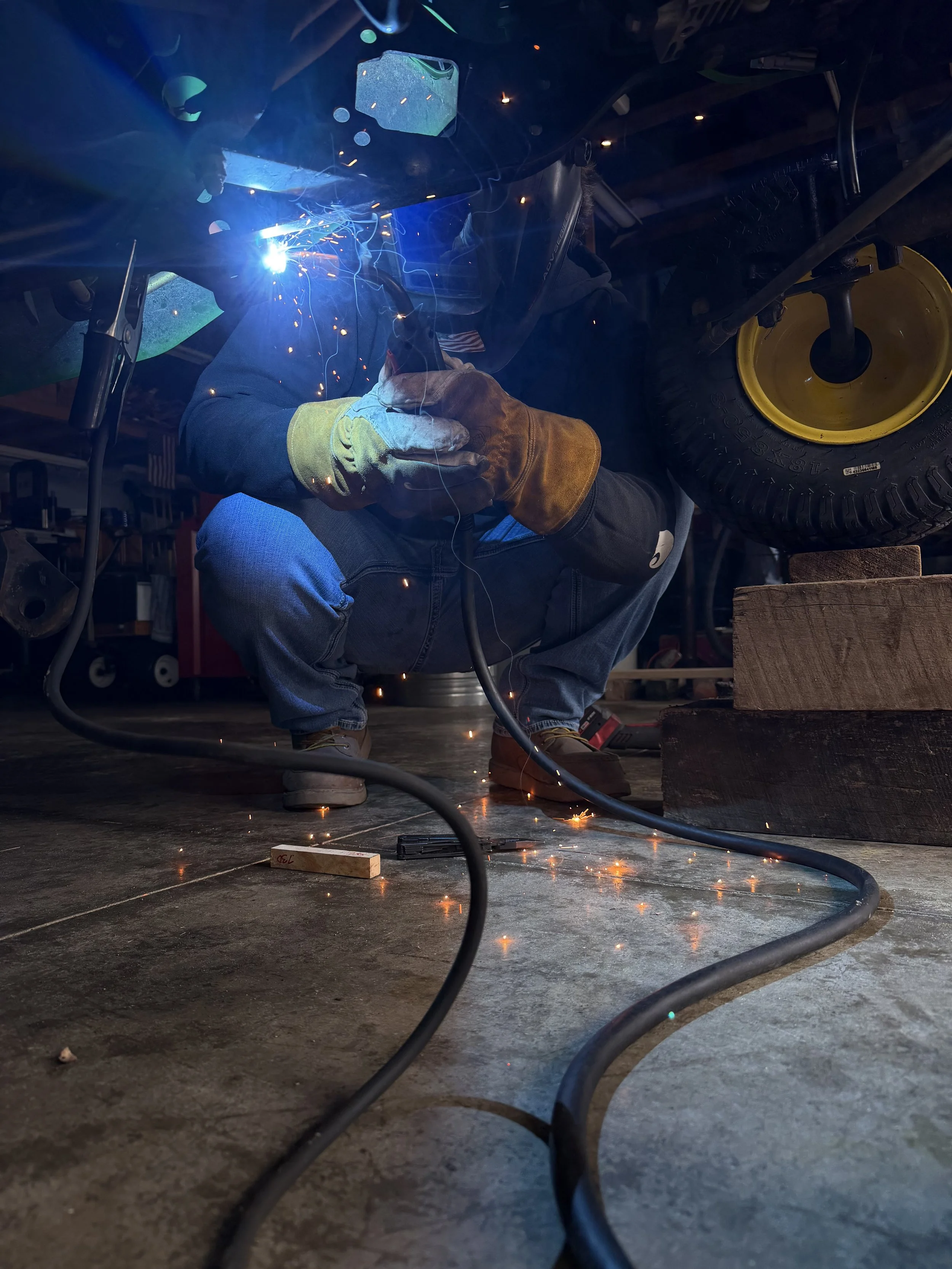 A person welding metal in a workshop, wearing protective gloves and a welding helmet, with bright sparks and blue light from the welding process visible.