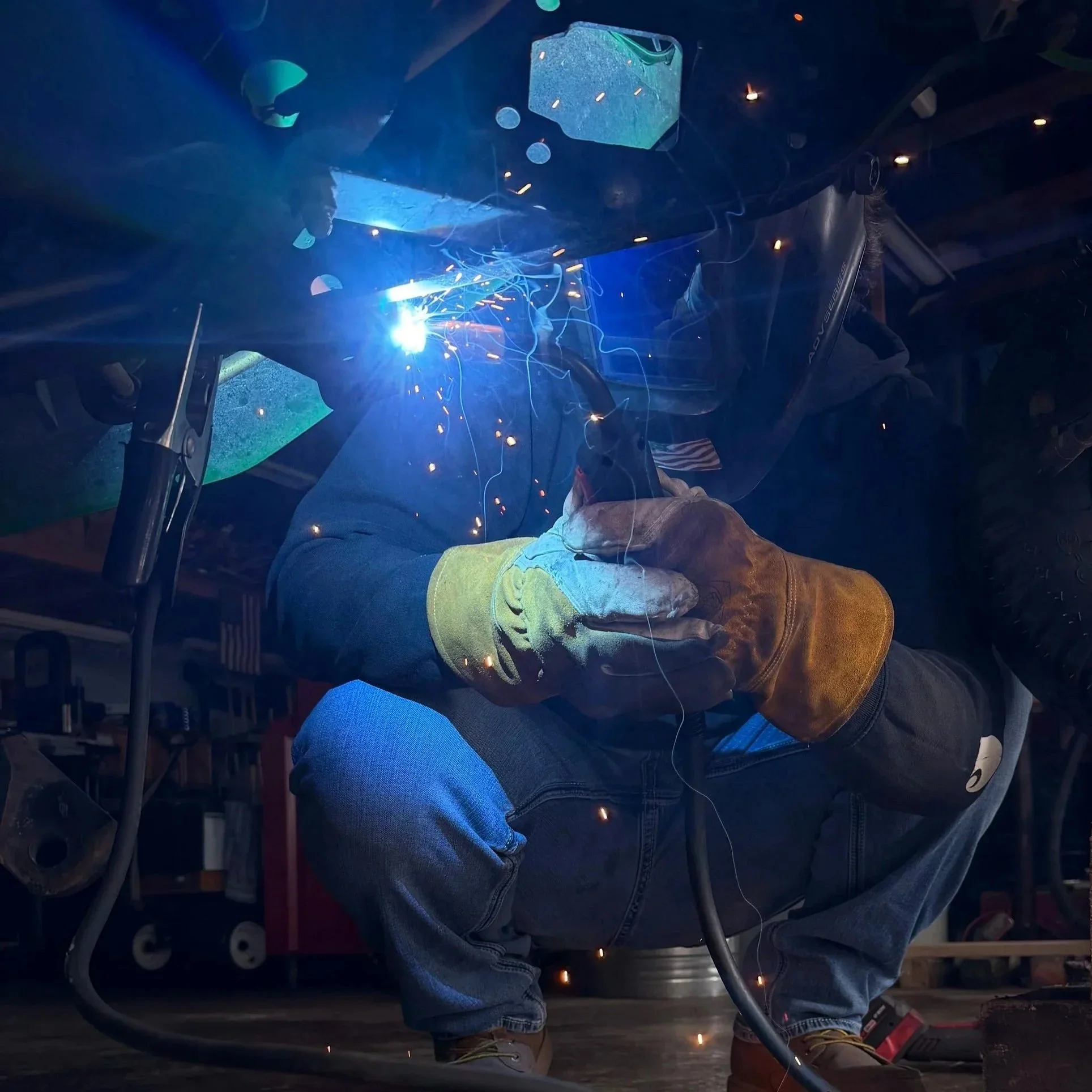 Technician welding with sparks flying while working on a metal structure in a workshop, wearing safety gear including gloves and a face shield.