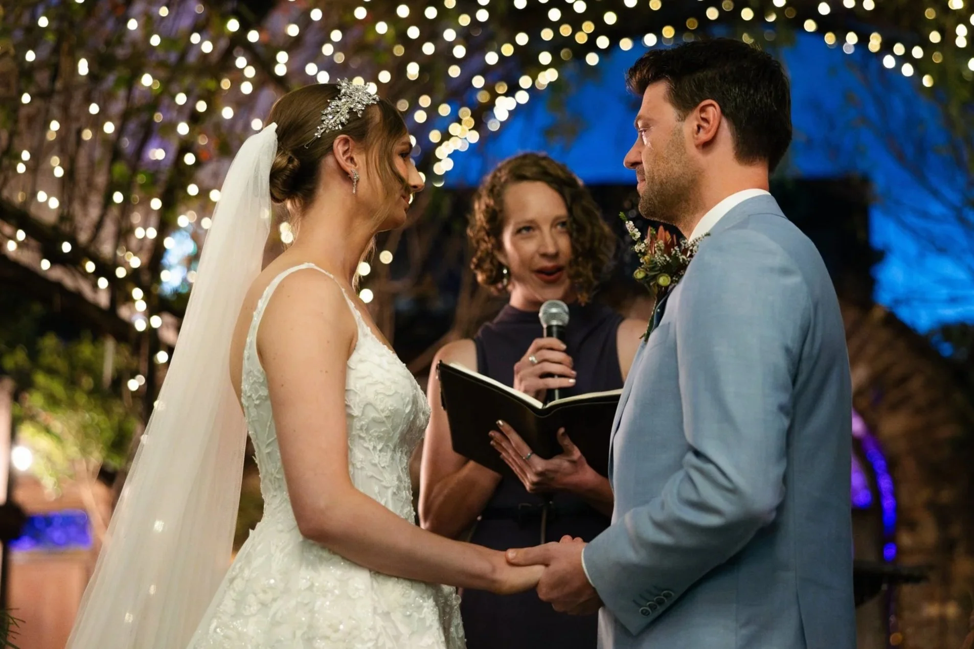 A bride and groom holding hands during their wedding ceremony, with a female officiant reading vows in front of string lights and a decorated tree.