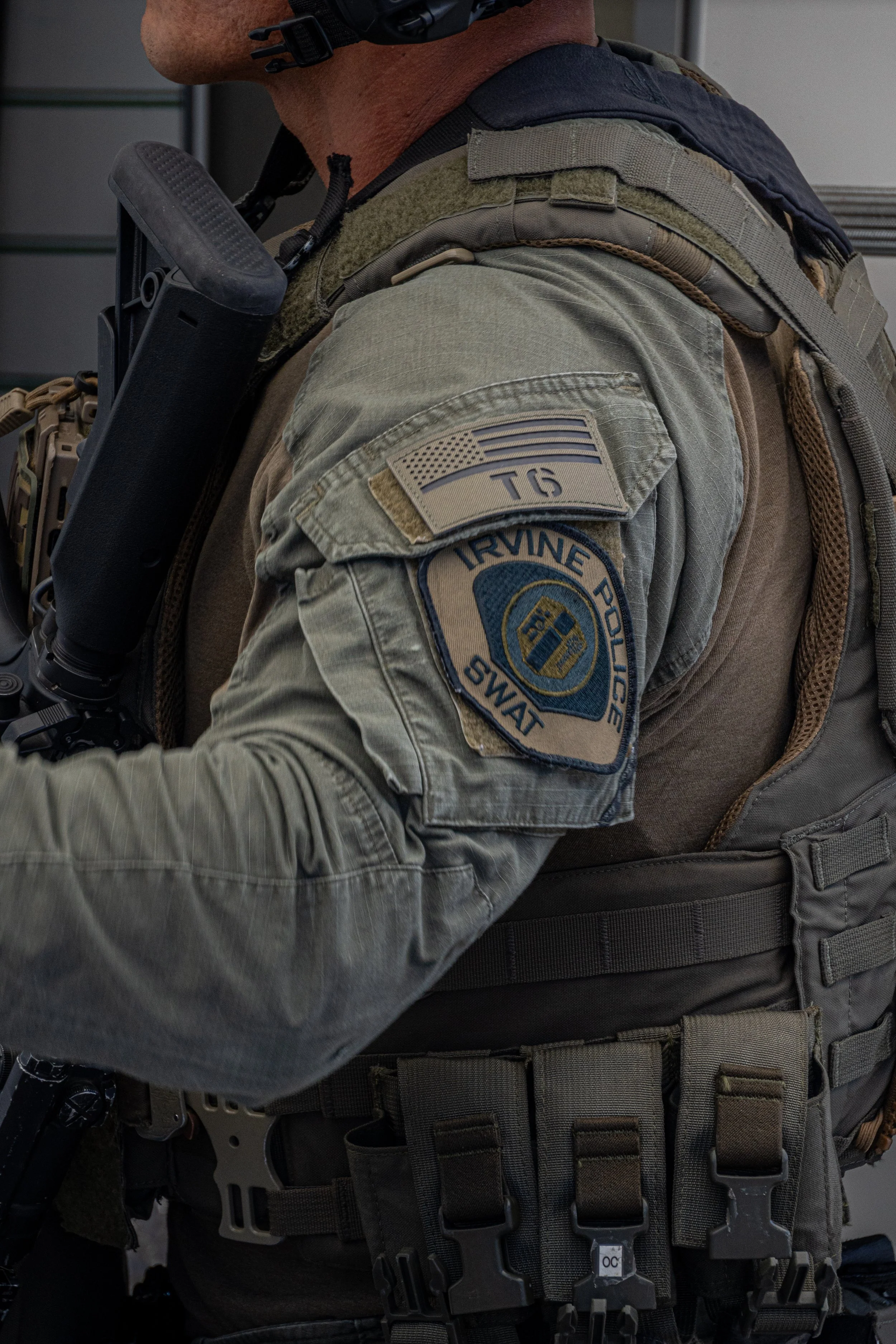 Close-up of a police officer's shoulder patch, which reads 'IRVINE POLICE SWAT' with an American flag patch above it. The officer is dressed in tactical gear, including a vest and a firearm.