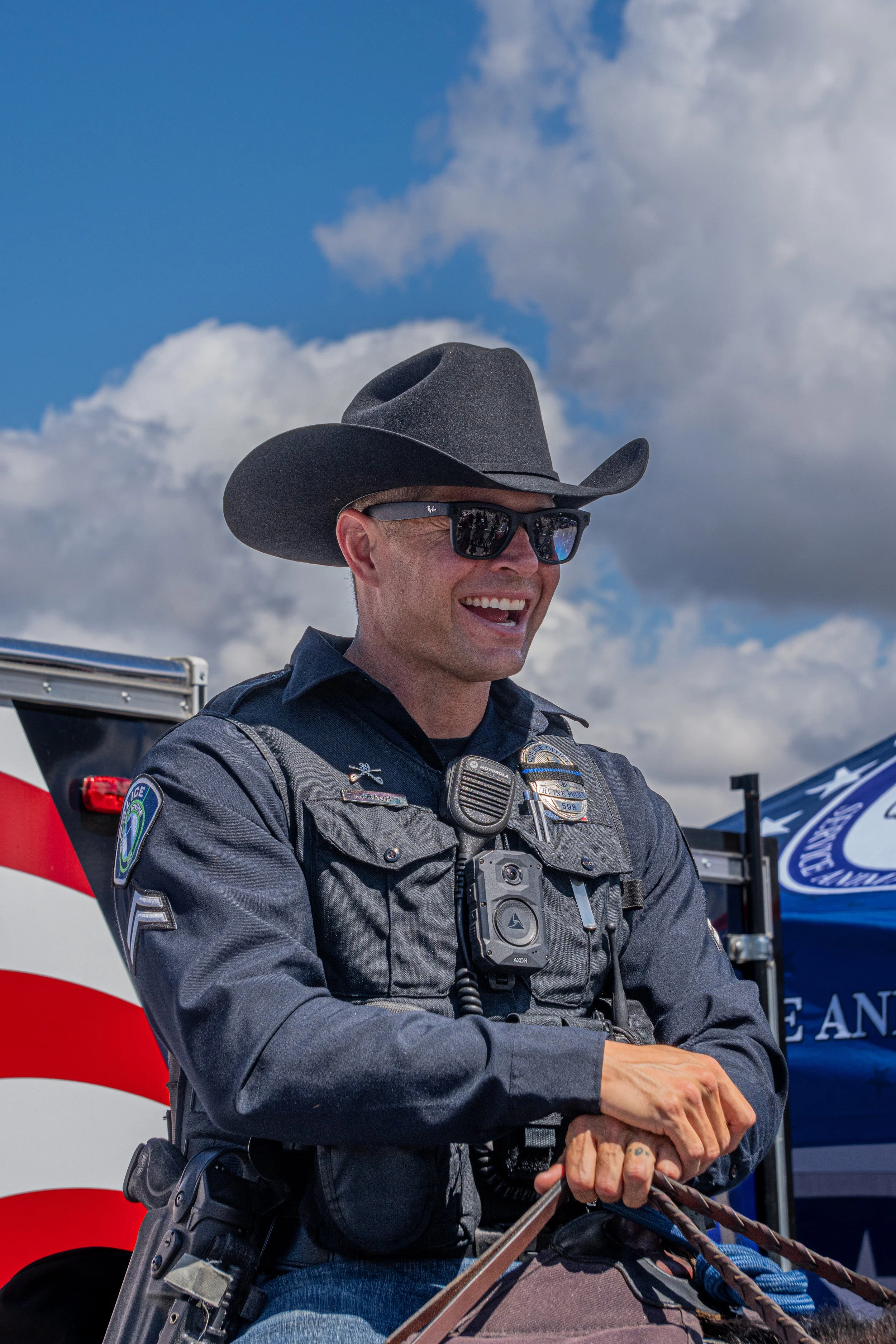 A smiling police officer wearing sunglasses and a cowboy hat, standing outdoors with blue skies and white clouds, with an emergency vehicle in the background.
