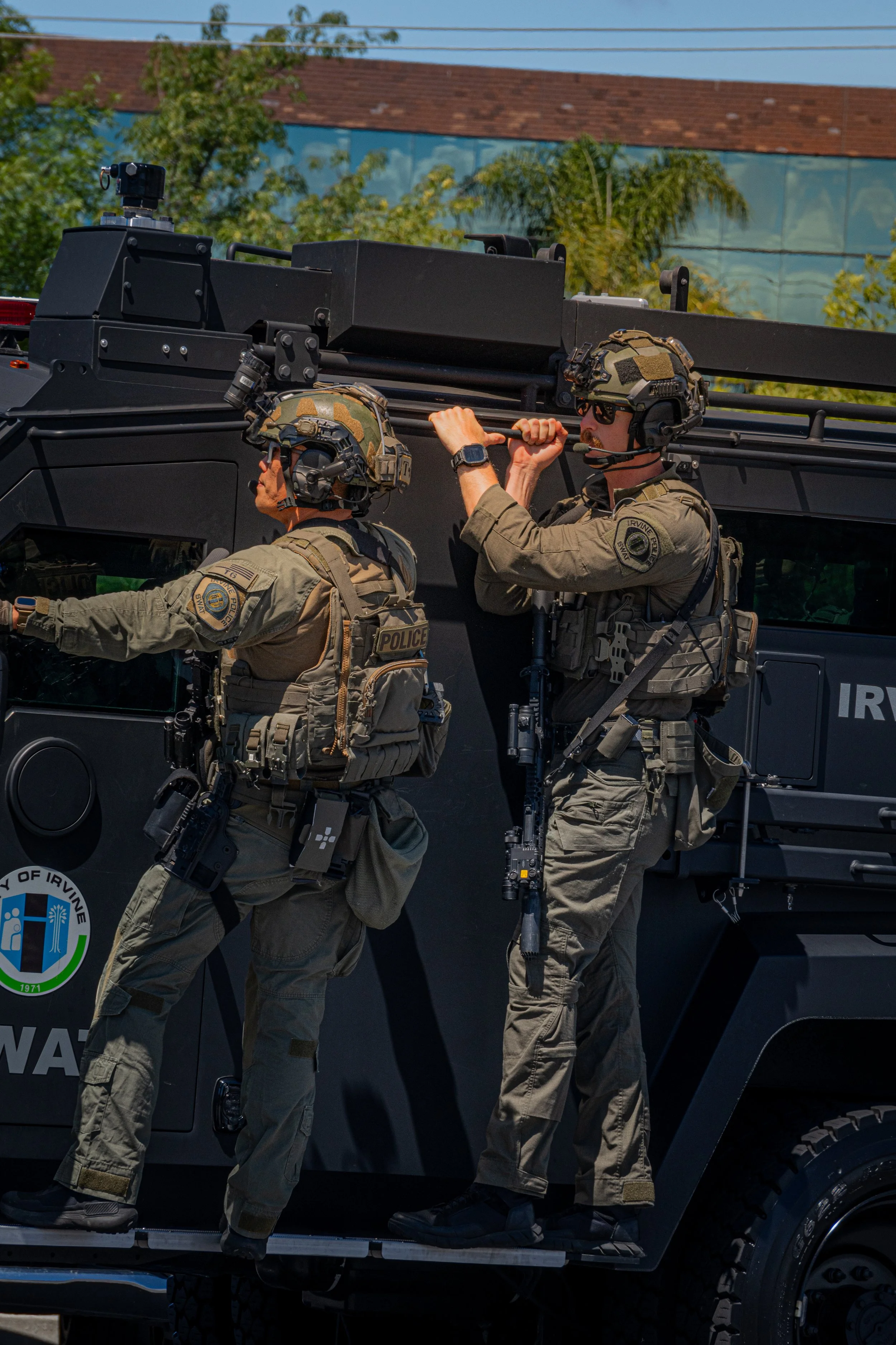 Two armed police officers in tactical gear on top of a black armored vehicle, with trees and a building in the background.