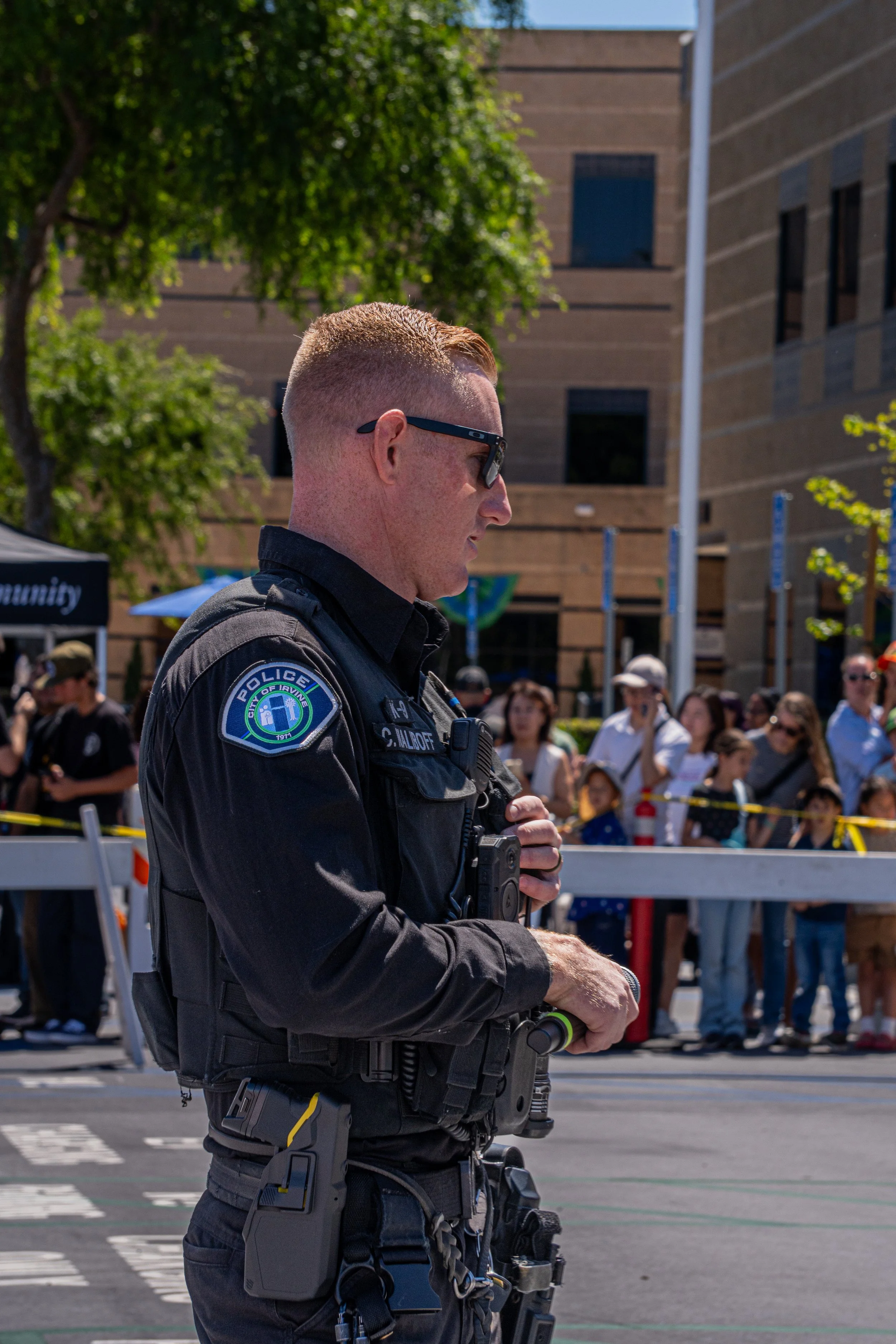 A police officer in uniform with sunglasses standing in a crowd at an outdoor event, holding a baton and wearing a radio.