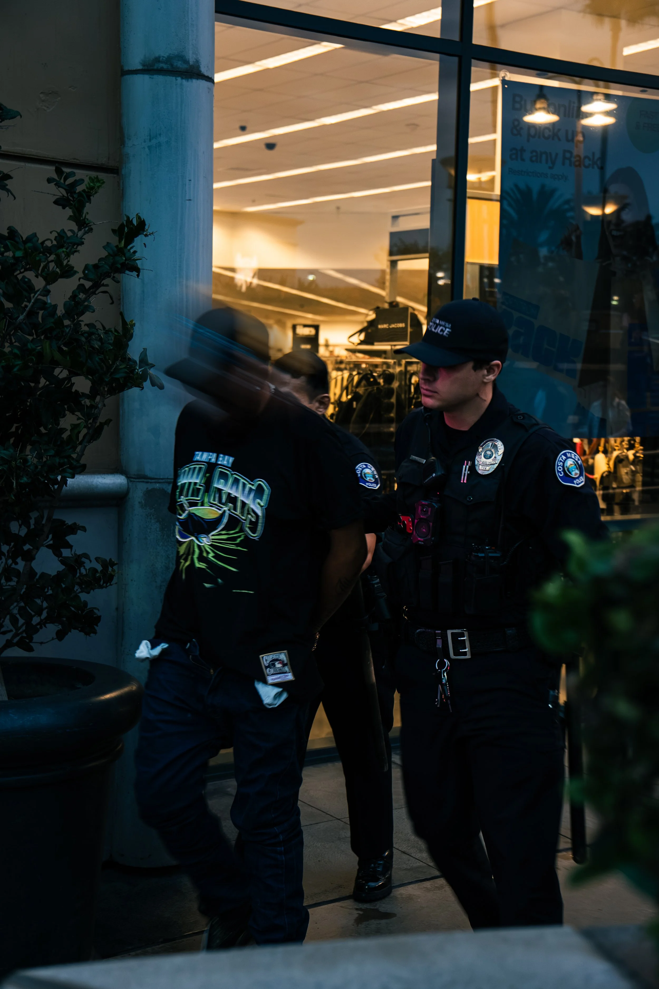 Two police officers are standing outside a building behind a glass window, engaged in conversation. One officer is in uniform with a badge and equipment, while the other is partially visible. The interior of the building appears to be a retail or gym