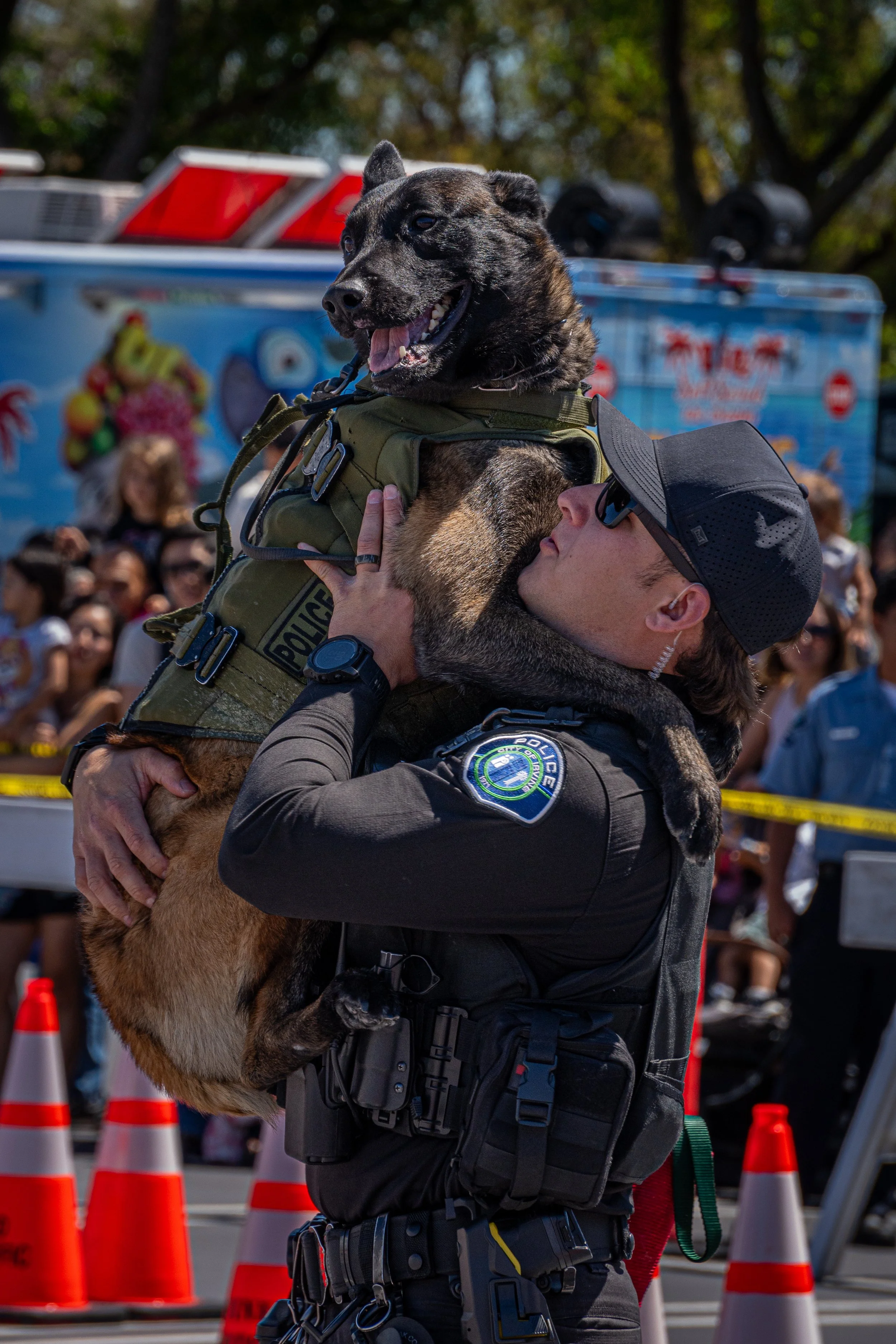A police officer hugging and kissing a police dog at an outdoor event with a crowd in the background.