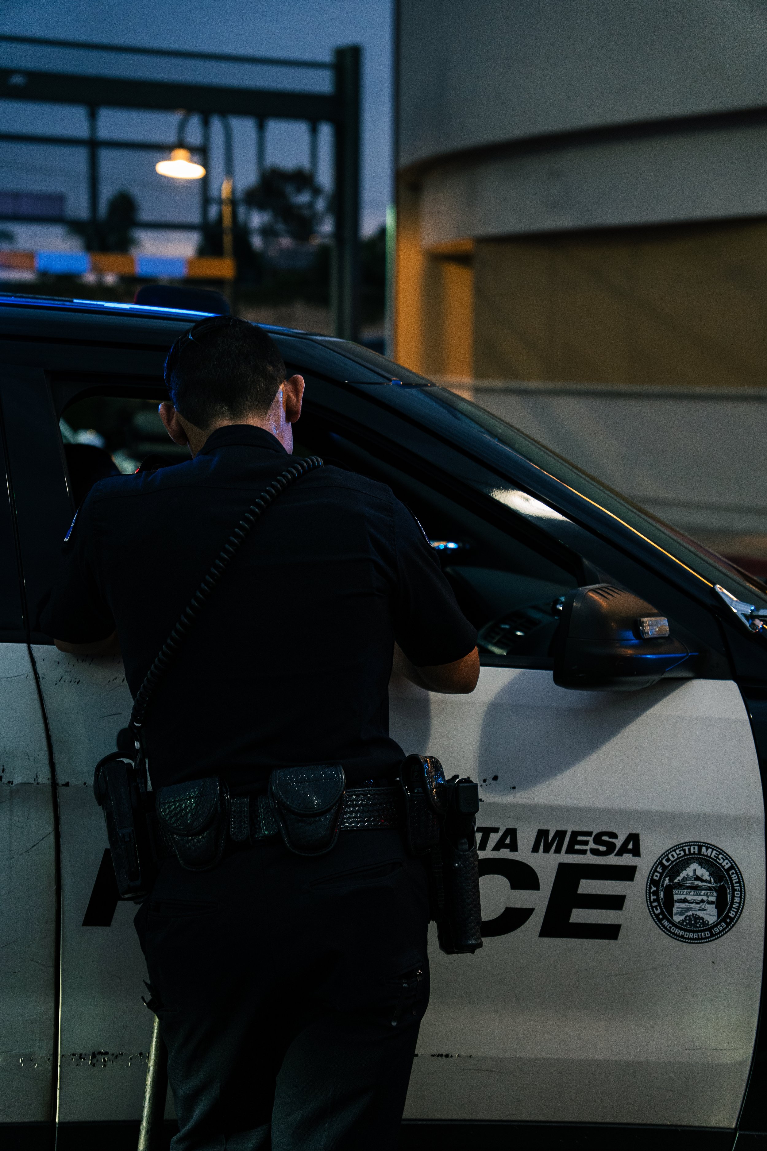 A police officer is writing a ticket or report in front of a police vehicle with the city seal of Costa Mesa, California. The scene is set during early evening or nighttime.